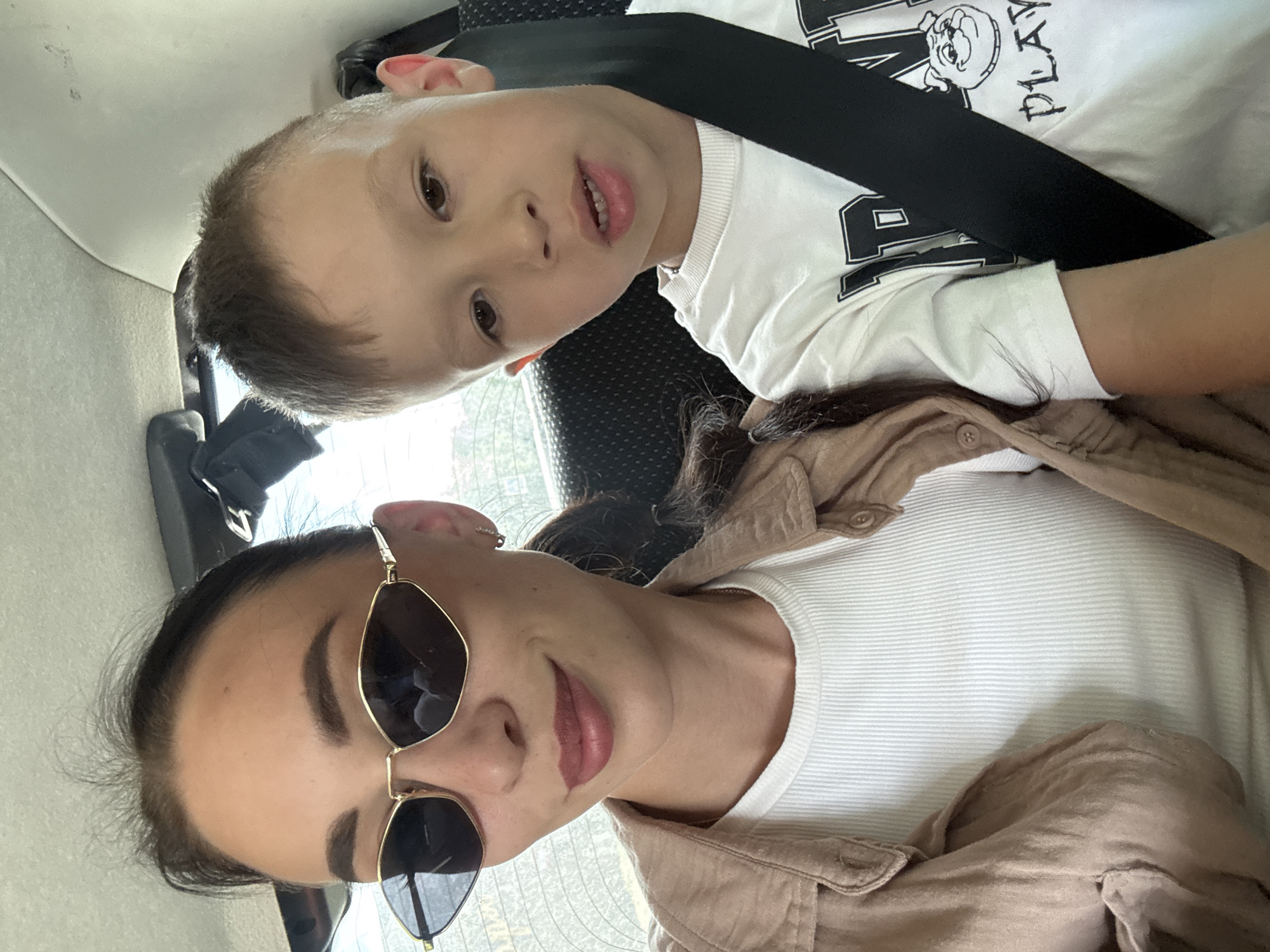 A selfie shows an adult and a child in what appears to be a car. The adult, a woman, wears sunglasses, a white ribbed shirt, and a tan jacket. The child, a young boy, wears a white t-shirt with a graphic design and is secured with a black seatbelt. The setting is suggested to be in the city of Chaikovsky, Russia, but there are no visual cues confirming this location. The photo is taken at an angle, capturing both faces.