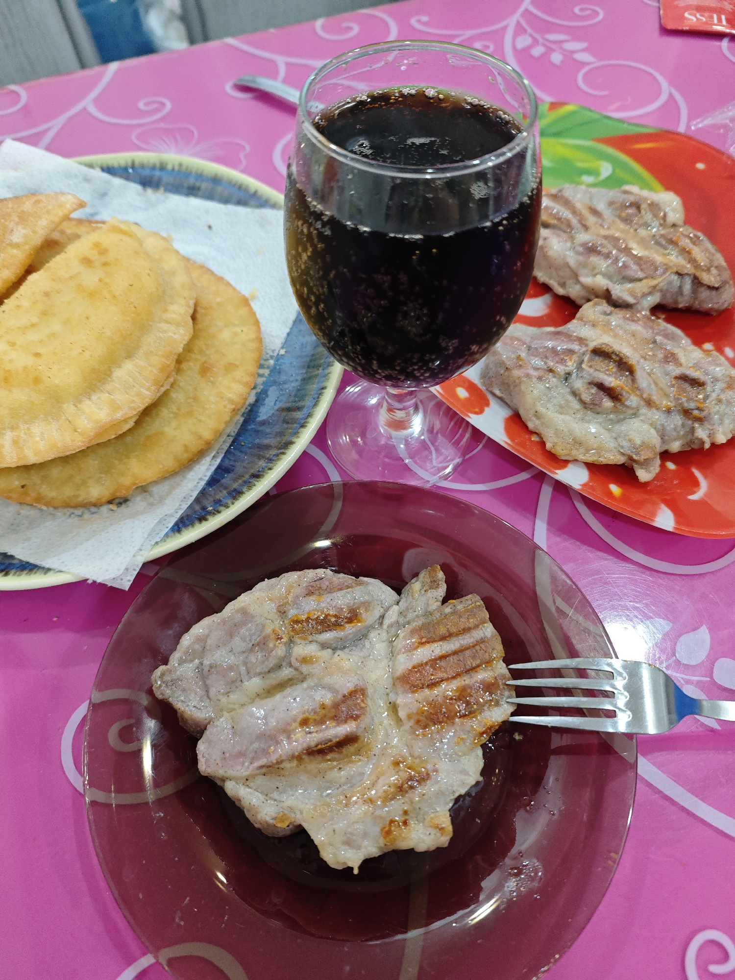 A high-angle shot depicts a meal on a pink tablecloth patterned with white floral designs. A red glass plate holds a grilled pork chop, pierced by a silver fork. Adjacent to it, a red plate with a floral pattern contains two more pieces of grilled pork. A blue plate with fried pastries is also present, along with a glass of dark soda. The scene appears to be set indoors, possibly in a private residence. The precise location within Markovskii, Russia, cannot be determined from the image alone.