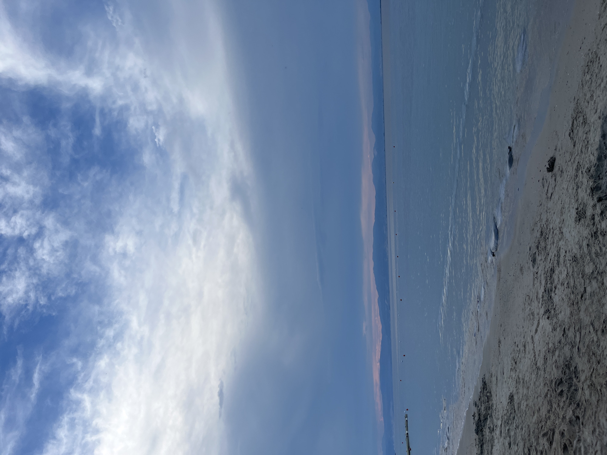 The image depicts a serene beach scene in Thassos, Greece, likely during the early morning or late evening. The foreground features a sandy beach with footprints, suggesting recent human activity. The gentle waves of the Aegean Sea lap the shore, creating a calm and tranquil atmosphere. In the distance, a range of mountains forms a picturesque backdrop against the horizon. The sky is a canvas of soft hues, with a mix of blue and white clouds, hinting at a changing weather pattern or the approaching dawn/dusk. Scattered buoys dot the water, indicating a designated swimming or boating area. The overall impression is one of natural beauty and peaceful solitude.
