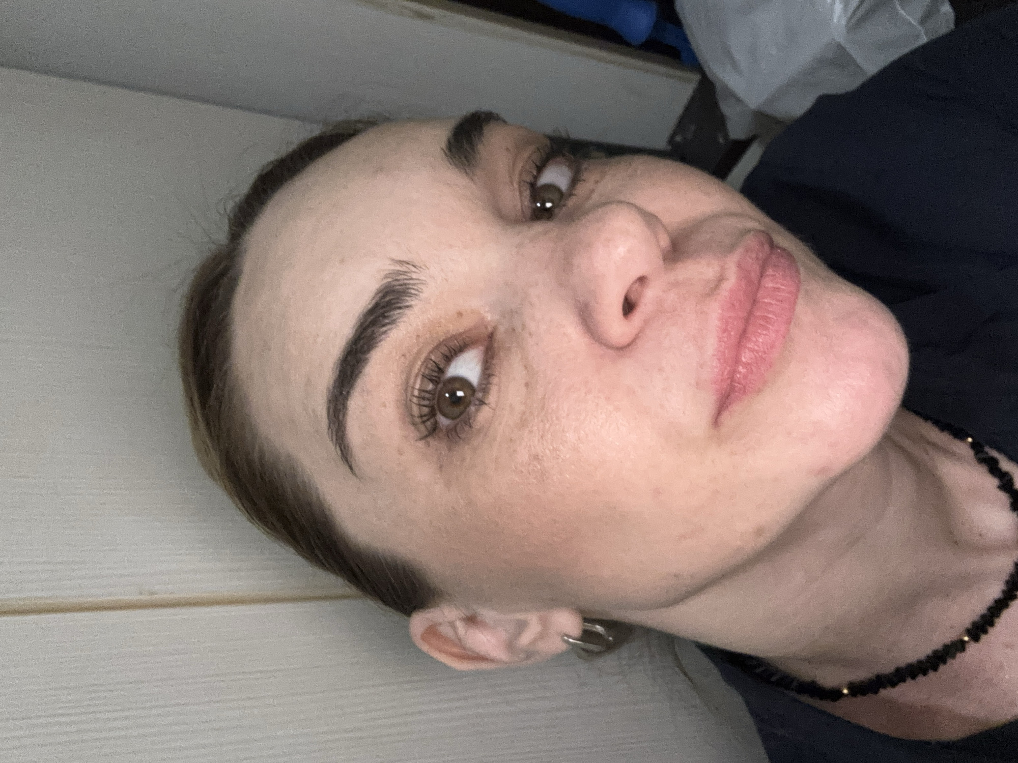 A close-up, angled self-portrait shows a woman with brown eyes and hair. She has fair skin with freckles and is wearing a black beaded necklace. She is wearing ear rings. The background includes a white wall and items out of focus. The image does not contain sufficient information to determine the location in Murino, Russia.