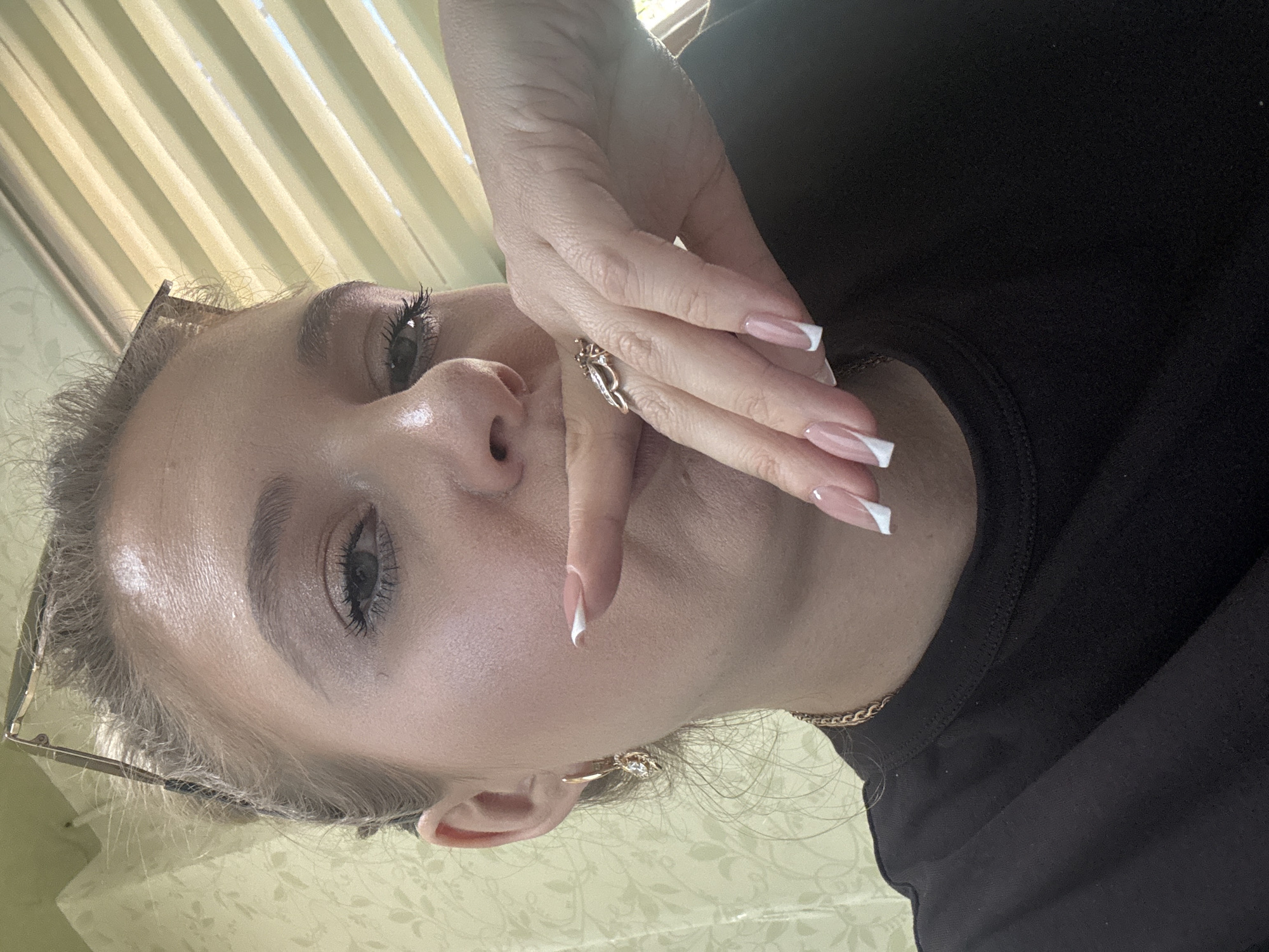 A medium shot captures a woman indoors, her face turned towards the camera as her right hand is raised to her mouth. She wears a black t-shirt and a gold chain necklace. Her nails are long and painted with a French manicure. She has on a ring on her right ring finger that appears to be butterfly-shaped, and a gold earring in her left ear. Her hair is pulled back, and a pair of glasses rests on top of her head. The background features a patterned wallpaper and vertical blinds.