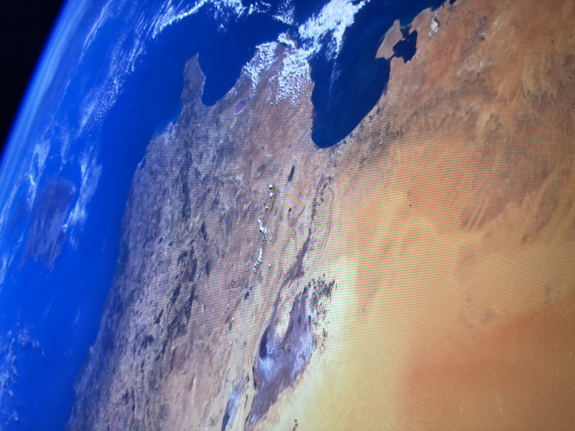 The image is an aerial view of a desert landscape with a large body of water. The land is predominantly sandy and brown, with some mountainous or hilly terrain visible. There are also patches of white, possibly snow or salt flats. The water is a deep blue and appears to be an ocean or large sea, with a coastline that is indented and features peninsulas. The sky is also blue, with some scattered clouds. The perspective suggests the image was taken from a high altitude, possibly from space or a very high-flying aircraft. There is no indication of the city of Kyiv, Ukraine, in this image.