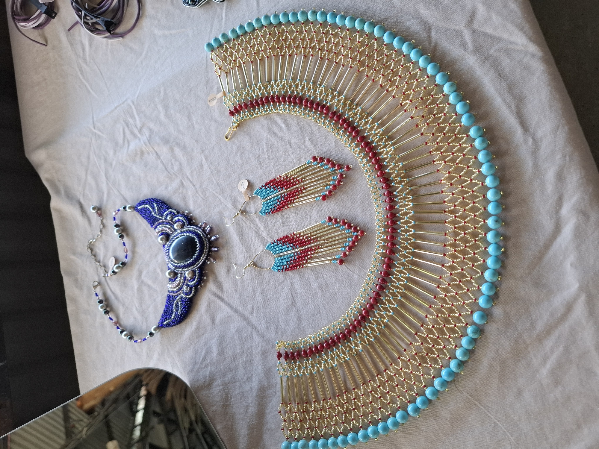 The image shows a collection of handmade jewelry laid out on a light-colored fabric surface. In the upper left quadrant, a partially visible mirrored surface reflects a blurred interior scene. To the right of the mirror is a blue beaded necklace with a large, dark oval stone centerpiece, accented with silver and pearl-like beads. Below and to the right of this necklace are a pair of matching earrings with red and blue bead fringes hanging from gold-colored rods. Dominating the lower portion of the image is a large, ornate gold-colored necklace featuring a wide, fan-like design. This necklace is adorned with rows of red and light blue beads, and has a fringe of numerous gold-colored rods, each tipped with a large, bright turquoise bead. The fabric surface appears to be a tablecloth or draped material. Two sets of coiled purple and white cords with clips are visible in the upper right corner. There is no indication of the location in Nha Trang, Vietnam.
