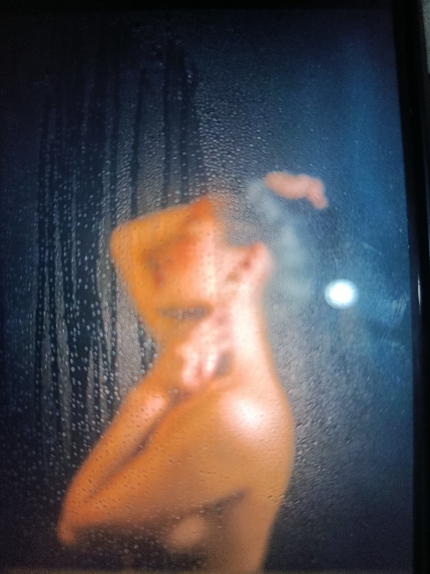 The image shows a blurry figure of a person through a steamy or wet surface. The person appears to be nude. There are water droplets on the surface through which the person is viewed. The background is dark. It is impossible to determine the location or city with the provided information.