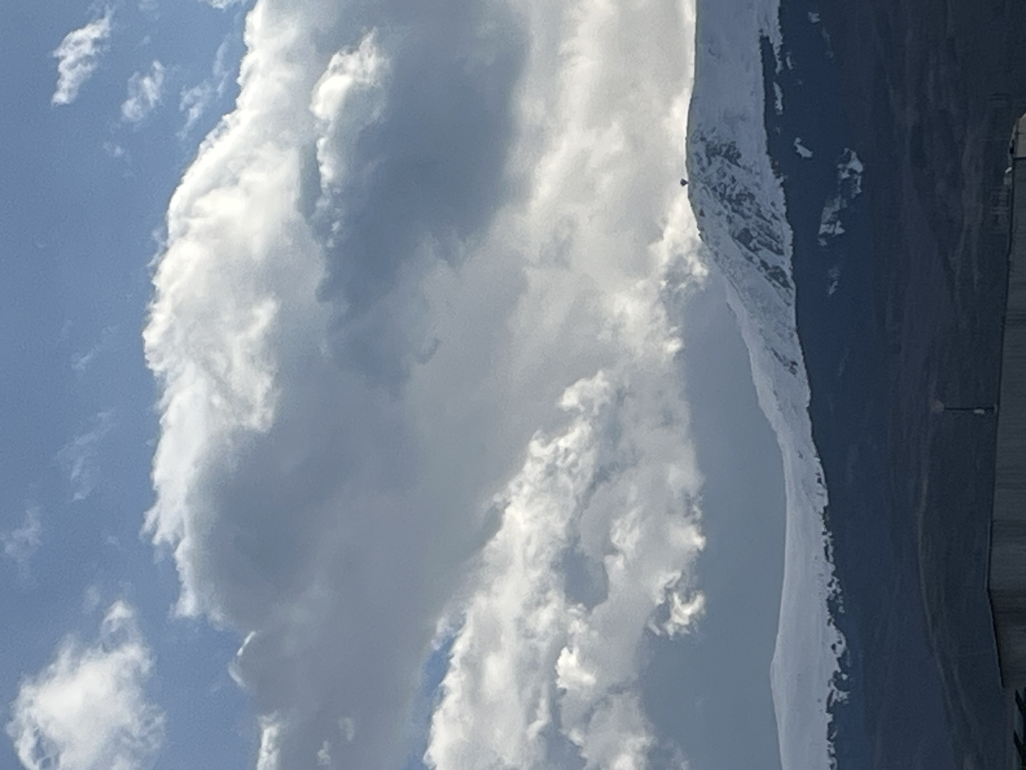 The image shows a landscape featuring clouds in the sky and a snowy mountain. The clouds are prominent, covering a significant portion of the upper half of the image. The mountain is located on the right side of the frame, with snow visible on its slopes. Part of a building is present on the lower-right corner of the image. The sky appears to be light blue in color where visible.
Location: Unknown, Unknown.