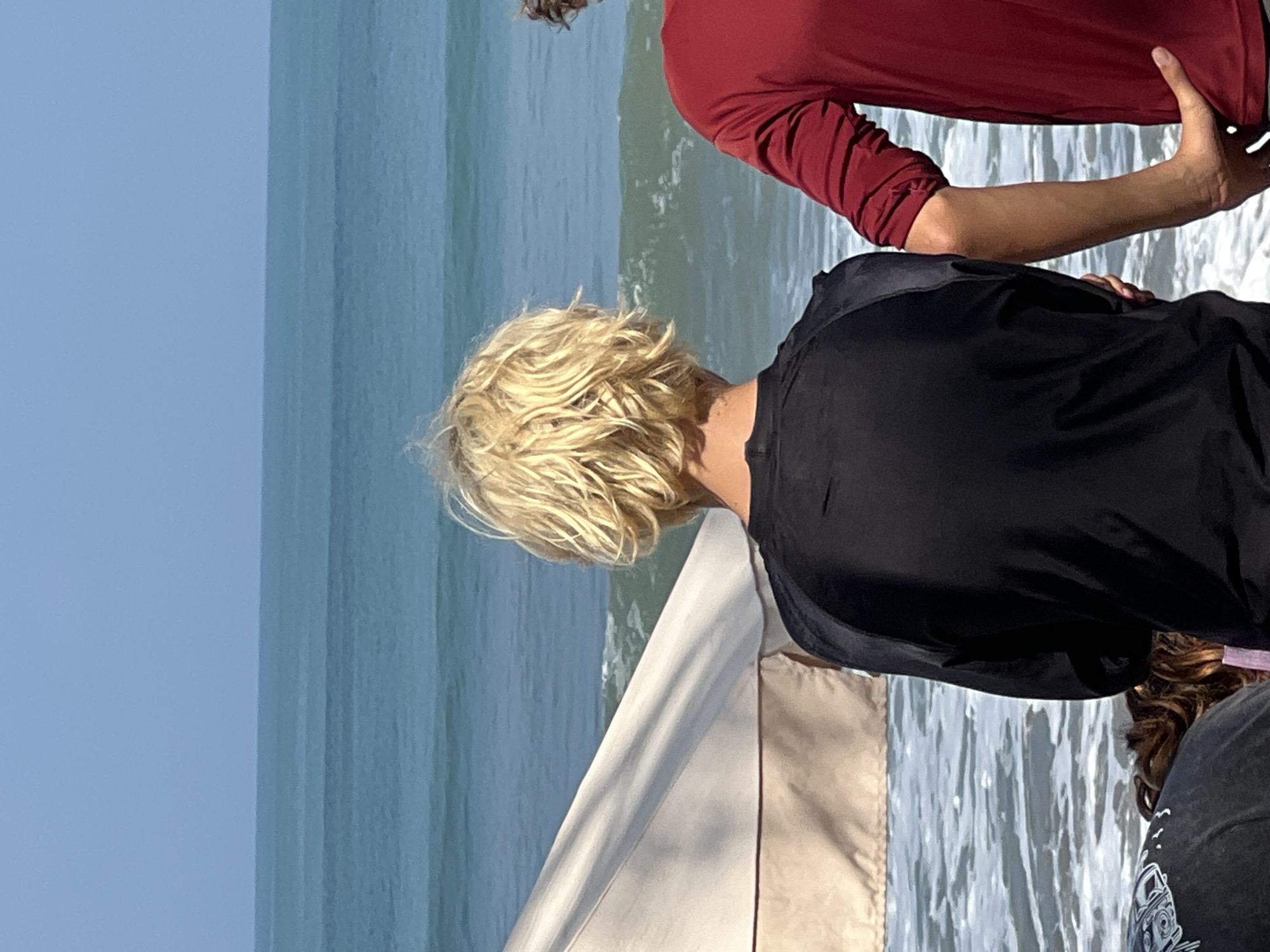 In Kuta, Indonesia, a group of individuals is gathered on a beach facing the ocean. In the foreground, a person with short, blonde, wavy hair wearing a black long-sleeved shirt is visible from behind, looking out at the sea. To their right, another person in a red long-sleeved shirt is standing, with their arm resting on the shoulder of the person in black. A light-colored beach umbrella is partially visible on the left side of the frame. The ocean is turquoise with white-capped waves, and the sky is a clear, light blue.
