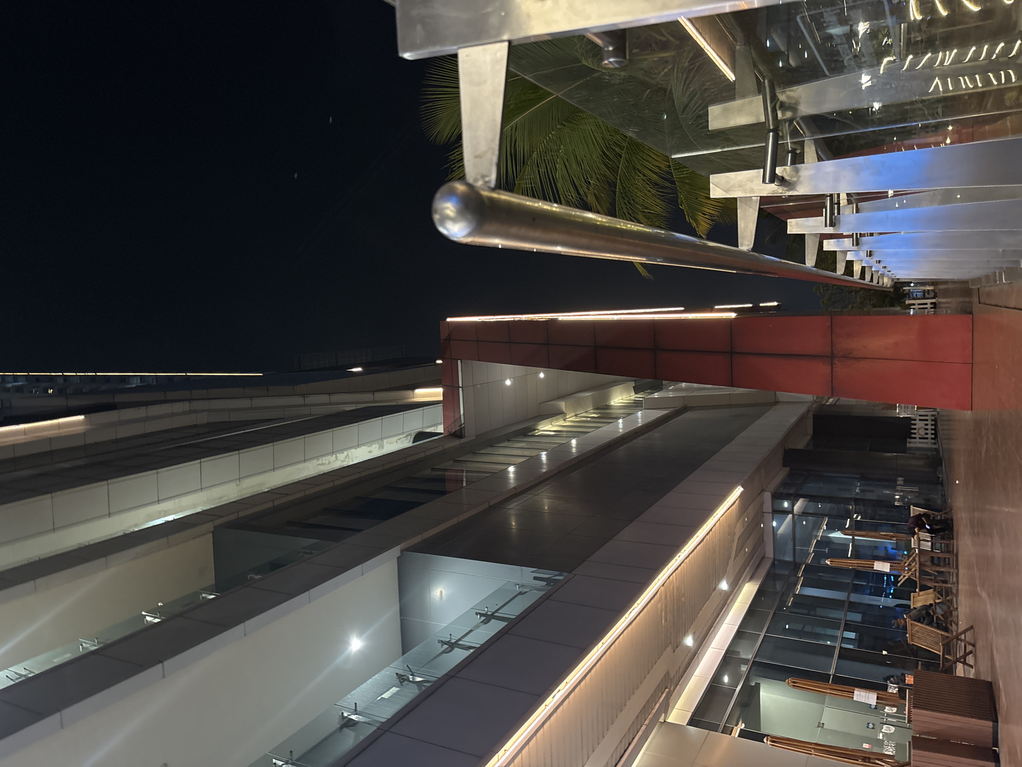 The photograph depicts a contemporary urban scene at night, likely in the city of North Jakarta, Indonesia. The perspective is from ground level, looking upwards at a multi-story building with a sleek, modern architectural design. The building features extensive glass facades, reflecting the interior lights and the dark night sky. Several stories are visible, with balconies and interior spaces illuminated.
A prominent red-colored vertical element is situated on the right side of the building's facade, adding a bold contrast to the otherwise muted tones of concrete and glass. Exterior lighting, in the form of warm-toned LED strips, runs along the edges of the building's various levels, highlighting its structure.
In the foreground on the right, a stainless steel railing and supporting posts create a visual barrier. Beyond this railing, glimpses of lush green palm fronds are visible against the dark sky, suggesting the presence of tropical vegetation, consistent with a location in North Jakarta.
On the left side of the image, at ground level, an outdoor seating area is set up. Several wooden chairs and tables are arranged, with some occupied by individuals. The area appears to be a cafe or restaurant with umbrellas shielding some of the tables. The ground surface is a smooth, polished material, possibly stone or concrete.
The overall atmosphere is one of a bustling yet serene urban environment at night, with a focus on modern architecture and outdoor leisure spaces. The lighting is artificial, emanating from the building's interior and exterior fixtures, creating a dynamic interplay of light and shadow.