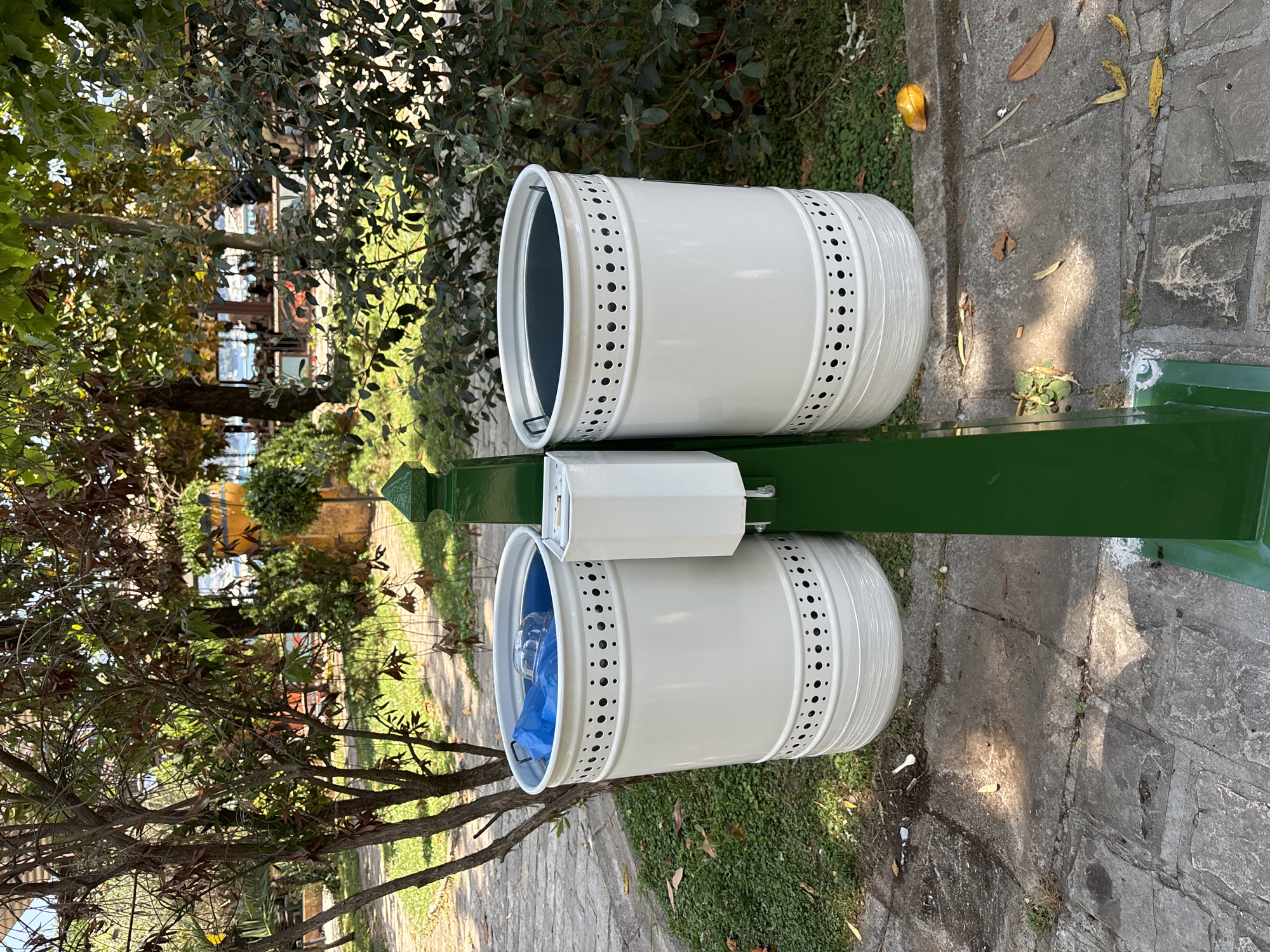 This is a medium shot, eye-level, outdoor photograph of a pair of white trash receptacles situated on a paved pathway in Budva, Montenegro. The receptacles are cylindrical with decorative perforated bands near the top and bottom. The receptacle on the left contains a blue trash bag, and the one on the right is empty. A smaller white dispenser, possibly for tissues or waste bags, is attached to the green metal pole that separates the two receptacles. The ground around the receptacles is a mixture of stone paving and grass, with fallen leaves scattered about. Behind the trash receptacles, trees and dense green foliage are visible. In the background, beyond the foliage, a body of water, possibly the Adriatic Sea, and distant buildings or structures can be seen. The lighting suggests a sunny day with shadows cast on the ground.