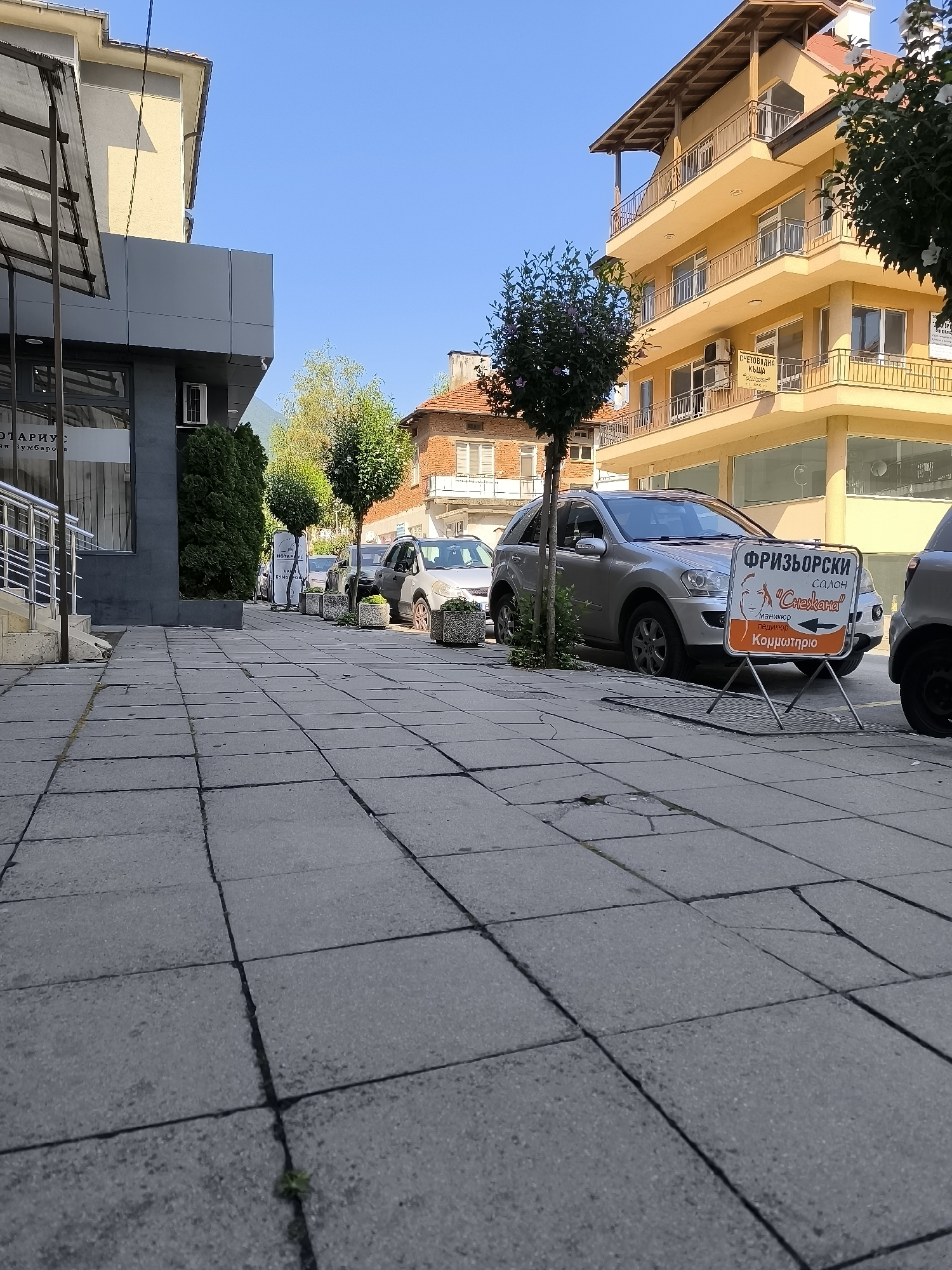 An eye-level, medium shot captures a street scene in Gotse Delchev, Bulgaria, on a clear, sunny day. The foreground is dominated by a grey, paved sidewalk with visible cracks. To the left, the modern facade of a building with grey paneling and a glass entrance is visible. To the right, a yellow, multi-story building with balconies stands prominently. In front of the yellow building, a silver SUV is parked parallel to the sidewalk. A brightly colored sign on a metal stand in front of the SUV advertises a "FRYZORSKI SALON" (hair salon) named "Snezana," indicating services like manicures and pedicures. Trees line the sidewalk, and other vehicles are parked further down the street. The background shows residential buildings and a hint of mountains under a clear blue sky. The overall impression is a quiet, everyday street scene in a Bulgarian town.