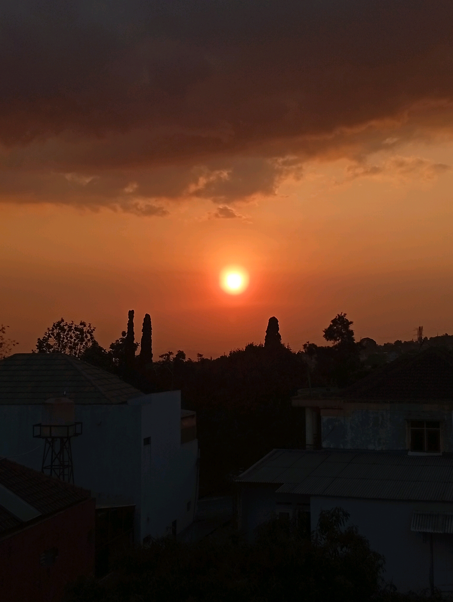 The image depicts a sunset over a residential area, likely in the city of Bandung, Indonesia, given the hint. The sky is a vibrant gradient of orange and dark brown, with a bright sun prominently featured near the center-right. Silhouetted against the sky are dark green trees and the distinct shapes of two tall, slender trees. In the foreground, several houses are visible, their rooftops and walls rendered in dark tones due to the low light. One house on the left has a water tank on a metal stand. The overall atmosphere is one of dusk in an urban or suburban setting.