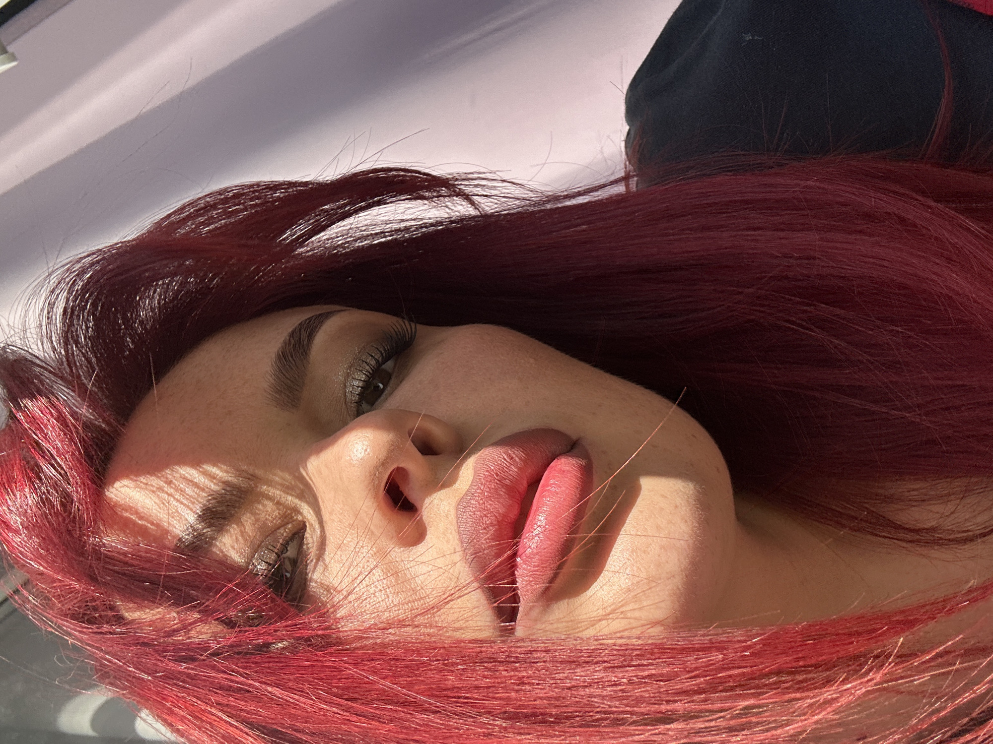 A close-up, low-angle shot captures a woman with vibrant red hair in Узюково, Russia. The sunlight casts dramatic shadows across her face, highlighting her features. She is wearing makeup, including eyeshadow and lipstick. Her hair, a striking red hue, is partially obscuring her face. The background is a soft, blurred purple.