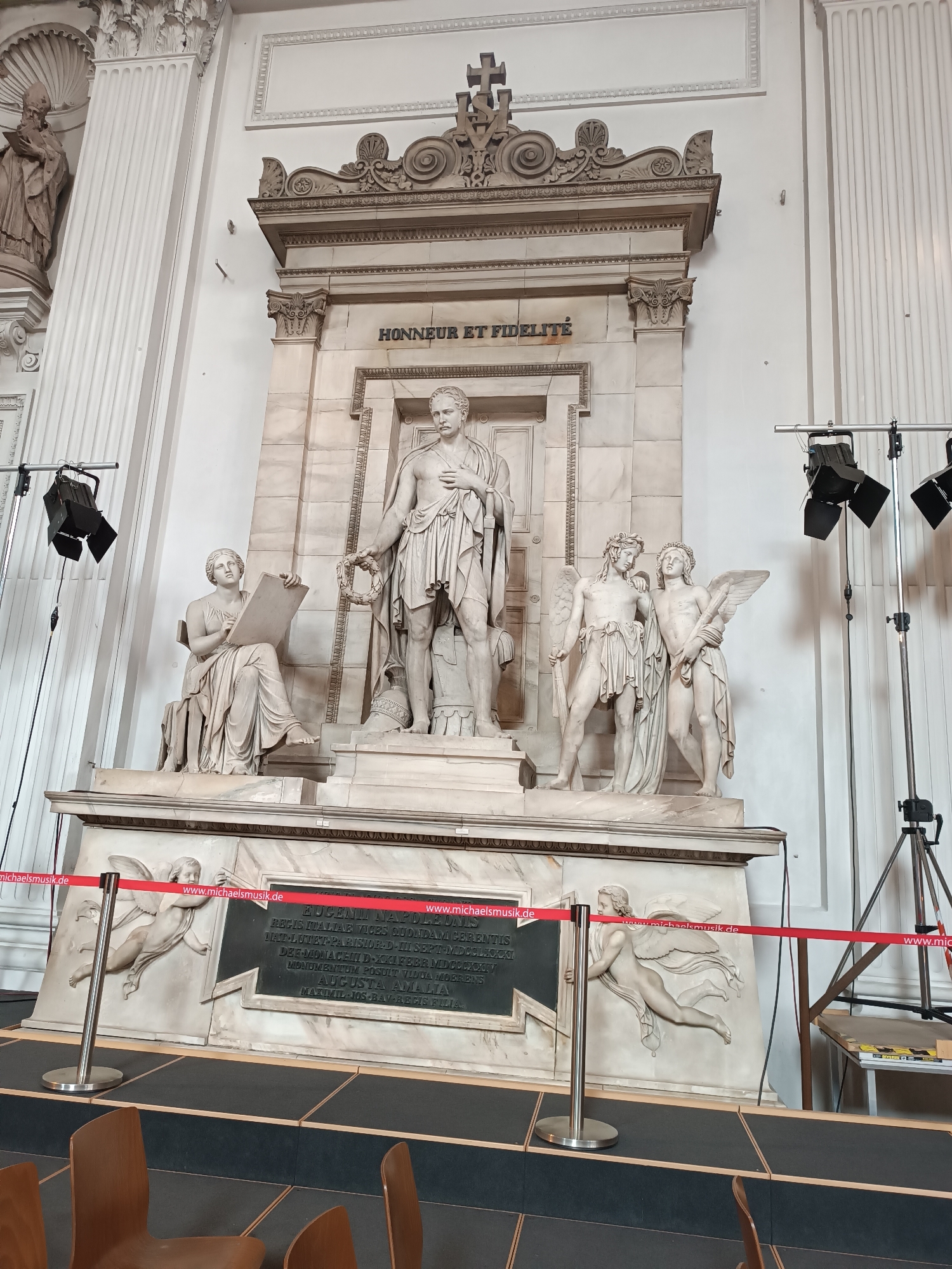 The image depicts a marble monument inside the Michaelskirche (St. Michael's Church) in Munich, Germany. The monument features several sculpted figures, including a central male figure standing with a wreath, a seated female figure to his left holding a tablet, and two winged cherubic figures to his right. The monument is topped with ornate carvings and an inscription that reads "HONNEUR ET FIDELITE". A red ribbon cordons off the area in front of the monument. The background shows architectural details of the church, including columns and sculpted reliefs. There are also lighting fixtures and stands positioned around the monument, suggesting it is being prepared for an event or exhibition.