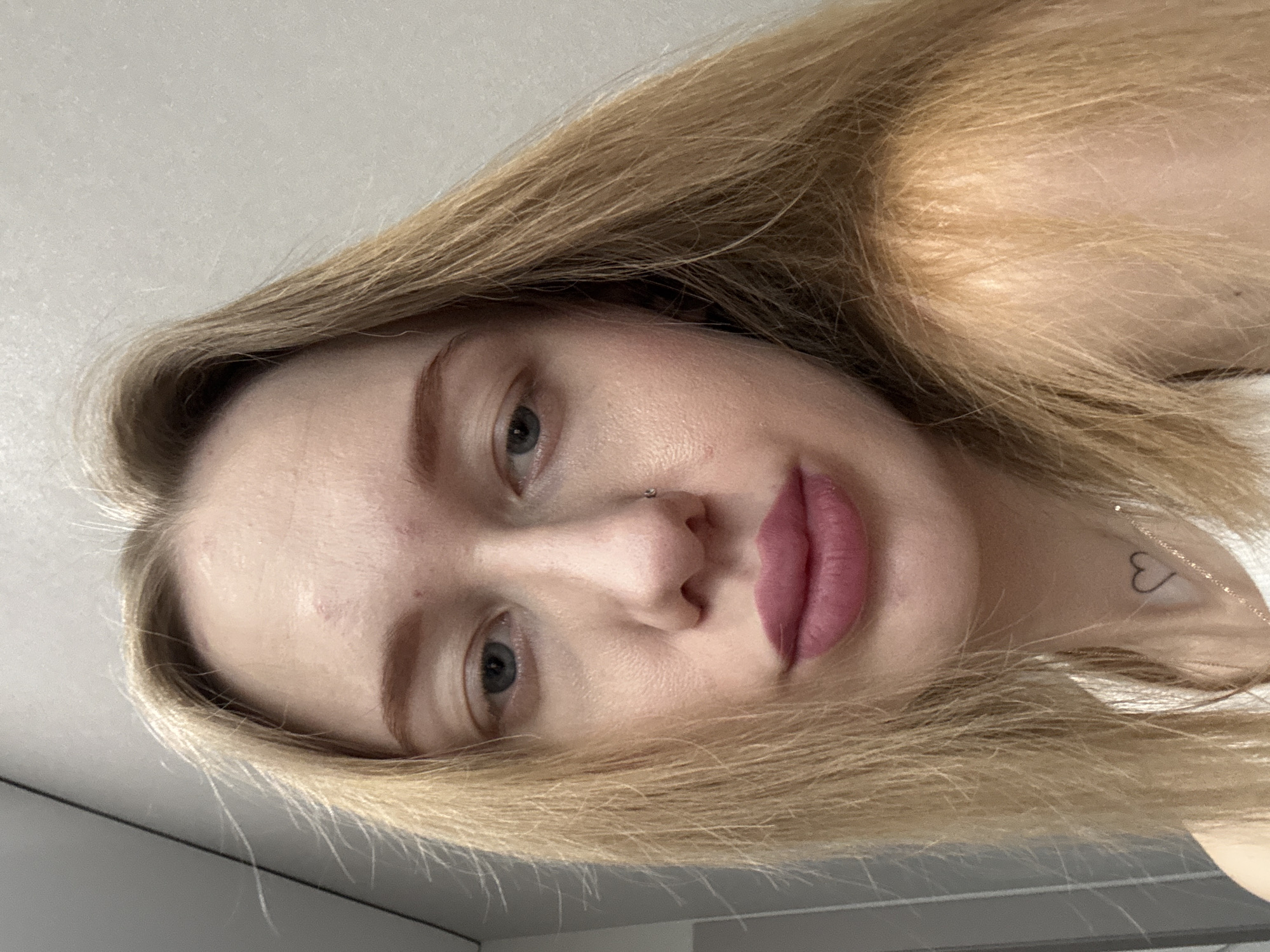A close-up, angled selfie shows a fair-skinned young woman with blonde hair. She has a nose piercing and pink lipstick. A small heart tattoo is visible on her neck. The background appears to be a plain, light-colored ceiling. The image doesn't have any indication of Ovoshchnoy, Russia.