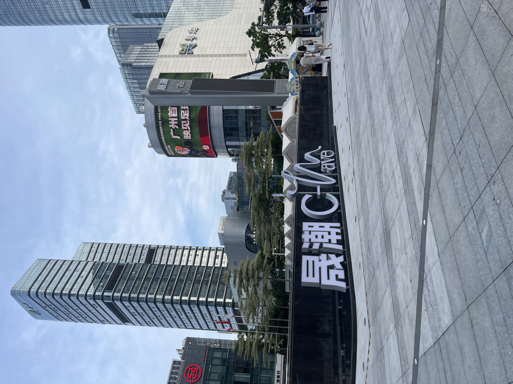 The image shows a modern urban scene in Panyu District, China, with a focus on a commercial area. Tall, sleek glass-fronted buildings dominate the background, reflecting the cloudy sky. In the foreground, a large, black sign with white Chinese characters and the English word "ave" is prominently displayed. The sign appears to be for a commercial establishment named "晟潮C".
To the left of the sign, a large digital billboard is visible, displaying colorful advertisements. One prominent ad features cartoon characters related to football, with Chinese text indicating "Guangzhou First" and "Watch Football". Another section of the billboard shows text related to "FIFA STO". Below this billboard, there are glimpses of a building with a prominent "天河城" (Tianhe City) logo, suggesting this is a shopping or commercial district.
Several people are visible in the scene, appearing to be pedestrians walking along a paved plaza. Some are casually dressed, suggesting it is daytime. The overall atmosphere is bustling and modern. The lighting indicates it is daytime, with overcast conditions. The presence of numerous commercial signs and modern architecture points to a developed urban environment. The image captures a snapshot of a busy commercial district in Panyu, China.