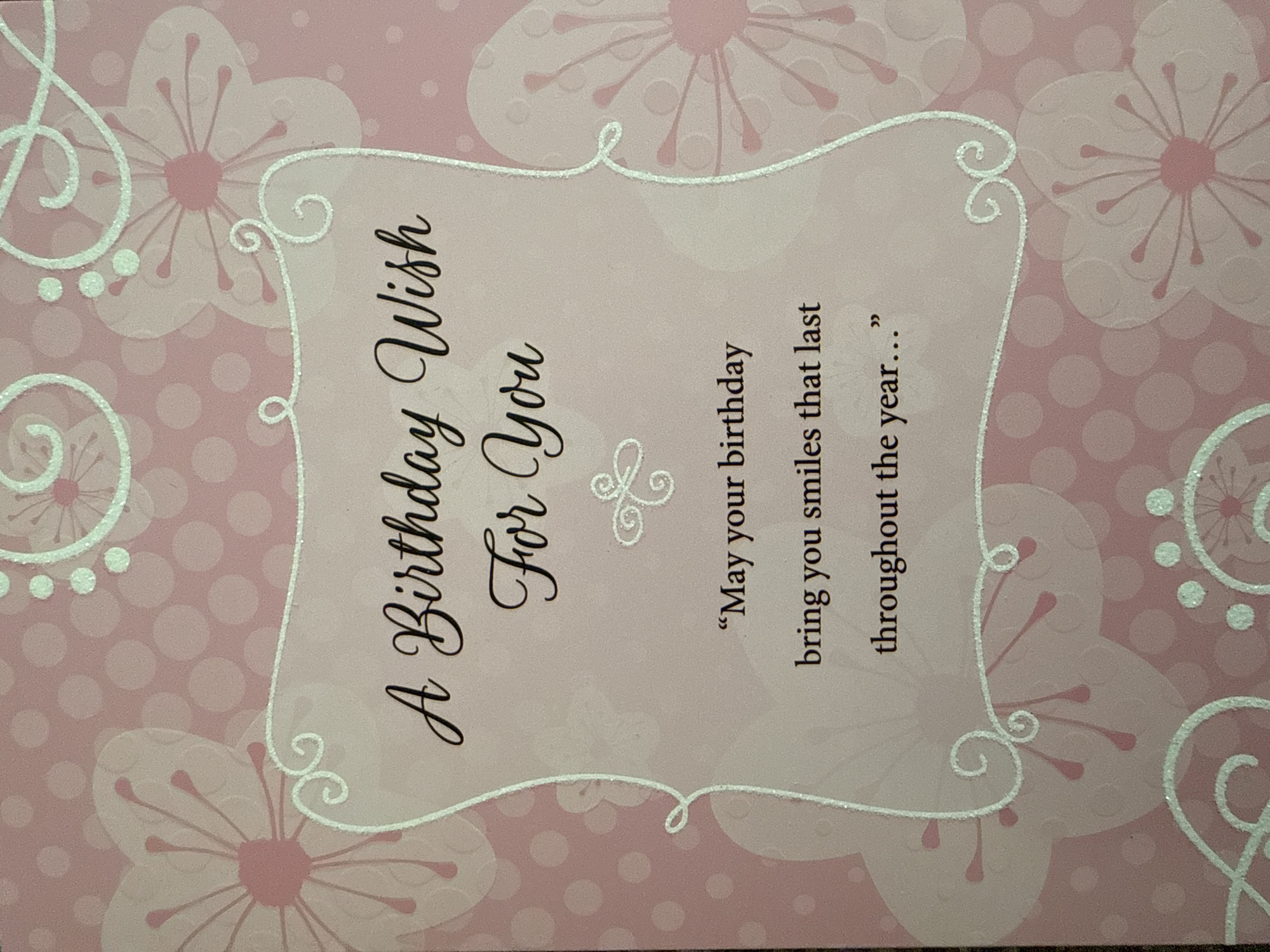 The image is a close-up of a pink birthday card with a floral design and glitter accents. The card features a message written in elegant script. The text on the card reads: "A Birthday Wish For You 'May your birthday bring you smiles that last throughout the year...'" The background of the card is a soft pink with a polka dot pattern and translucent floral illustrations. There are no people or specific locations in the city of West Sacramento visible in the image.