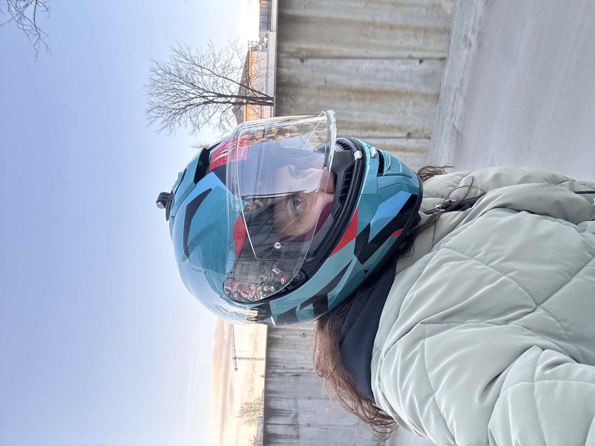 A selfie shows a person wearing a blue motorcycle helmet with the visor partially raised. The helmet has black, red, and white accents. The person is wearing a light green quilted jacket. The background is an urban scene with a concrete structure and a tree without leaves against a pale sky. A crane is visible in the distance. The image was taken in the city of Sofia, Bulgaria.