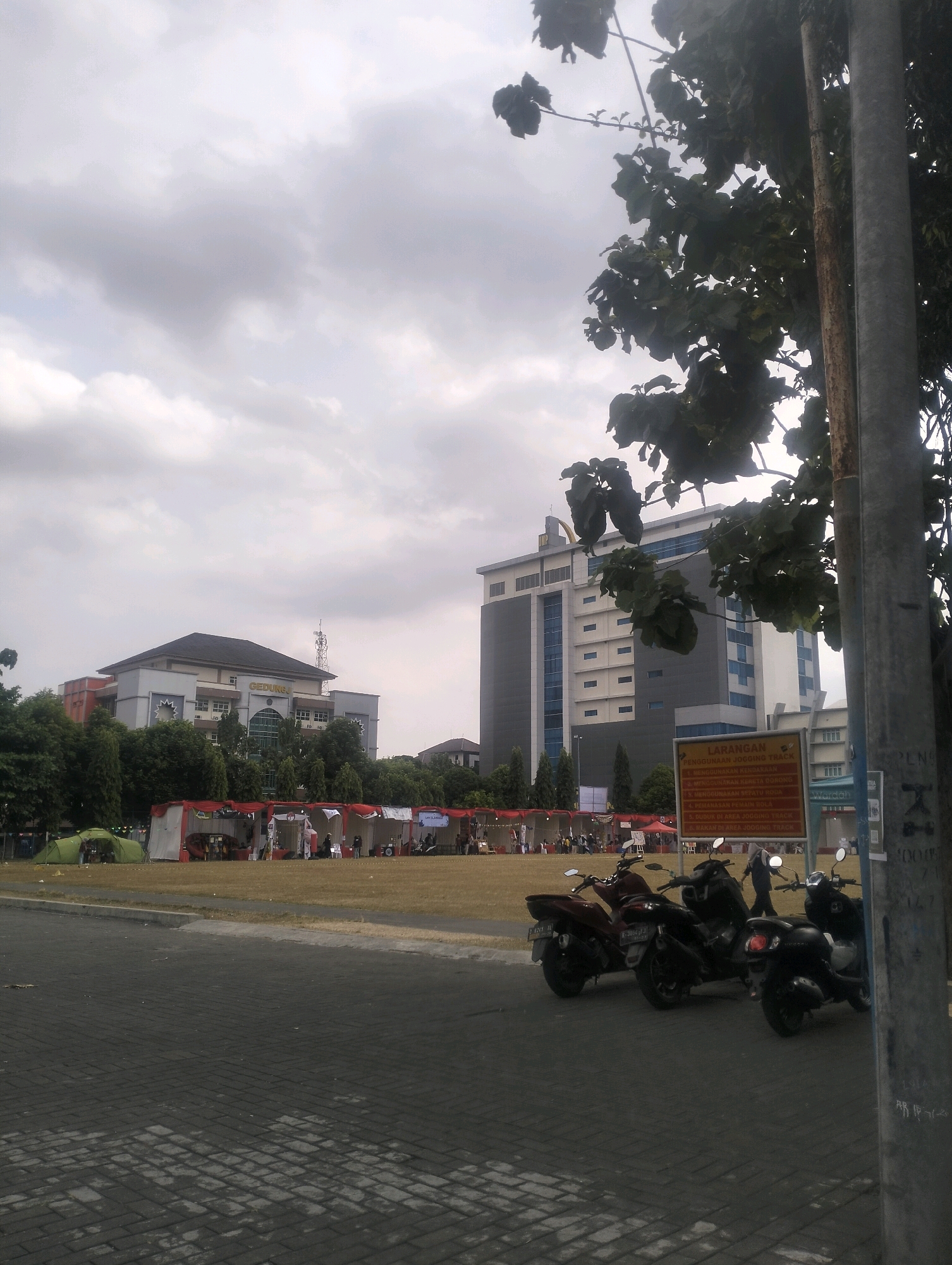 The image depicts an outdoor scene with several buildings and tents in the background. In the foreground, there are several motorcycles parked on a paved surface. A large tree with dark green leaves is visible on the right side of the frame, partially obscuring a metal pole. The sky is cloudy.
In the middle ground, there are numerous white tents with red accents, suggesting an event or market is taking place. Behind the tents, a building with the word "GEDUNG" visible on its facade stands. Further in the background, another taller, modern building with a gray and white exterior is present. A green tent is also visible on the left side of the field.
A red and yellow sign with Indonesian text is posted on a pole to the right of the tents. The text appears to be a list of regulations, likely related to the area or event. Some people are visible near the tents, and a few are standing or walking around.
The overall impression is that of a bustling outdoor event or gathering, possibly at a university campus or public space in Karangsoka, Indonesia, given the visual cues and the context provided. The weather appears overcast, suggesting a potentially humid climate.