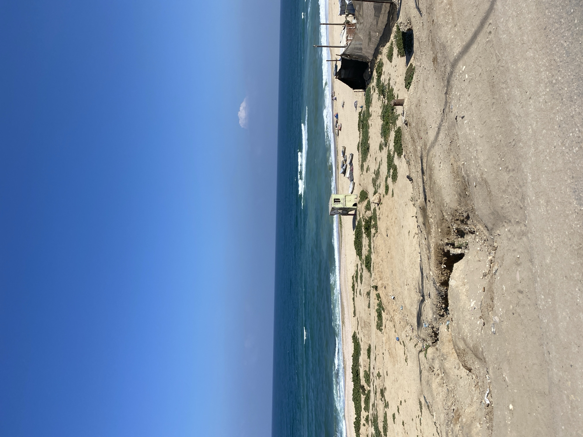 The image presents a seaside view, likely taken from an elevated position such as a bluff or high ground adjacent to the beach. The sea occupies the central part of the frame, with waves visible, hinting at moderate to significant wave action. To the left, a vast expanse of clear blue sky suggests a sunny day. On the right side of the image, there is a sandy terrain with sparse vegetation. A dirt or concrete path runs along the edge, showing signs of erosion and instability, particularly with the presence of a significant cavity or hole in the ground. This feature, along with the general texture of the ground, suggests potential infrastructural issues or environmental impact on the land. There are some indistinct structures near the beach and objects that seem to be small fishing boats.
