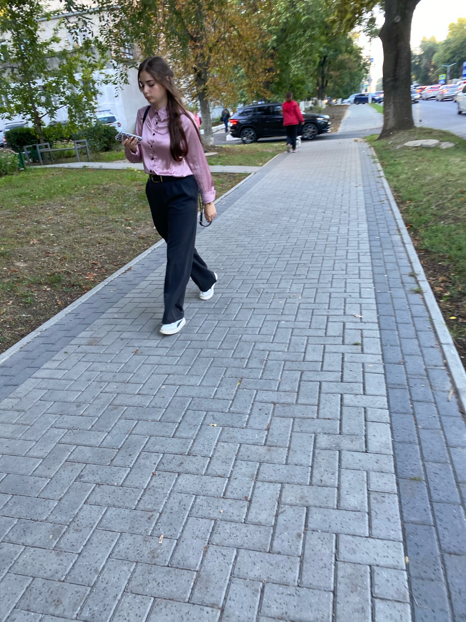 A young woman with long brown hair, wearing a pink silk blouse and black trousers, walks on a paved sidewalk, looking at her phone. She is wearing white sneakers. Behind her, a woman in a red jacket walks a small white dog. A black SUV is parked on the side of the road. The scene is in a city with trees lining the sidewalk and cars on the road in the background. The paving stones are arranged in a herringbone pattern. The overall impression is of an urban environment on a mild day.