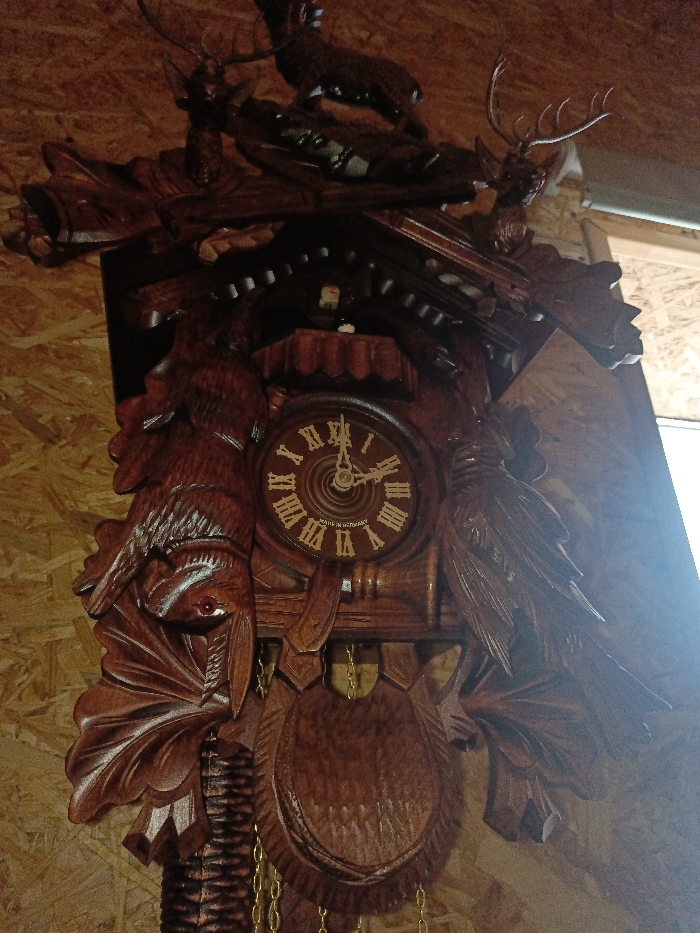 A large, intricately carved dark wooden cuckoo clock is depicted, mounted on a light brown wooden paneled wall in Gorka, Russia. The clock features detailed hunting-themed carvings, including two deer heads with antlers flanking the top, a deer figure positioned centrally above the clock face, and carved birds flanking the sides of the main clock mechanism. A carved rabbit or hare hangs inverted by its legs below the left bird carving, suggesting a hunting trophy. The circular clock face displays gold Roman numerals (I-XII) and two dark hands indicating approximately 02:45. The inscription "MADE IN GERMANY" is visible on the lower portion of the clock face. A small, arched door is situated above the clock face, with a white cuckoo figure partially visible inside. Below the clock face, a carved wooden pendulum, designed as a hunting shield or animal head, is suspended, with two golden chains extending downwards, presumably connected to weights. The scene is illuminated by a strong, unseen light source to the right, creating significant glare on the right side of the clock and the wall. No individuals are present.