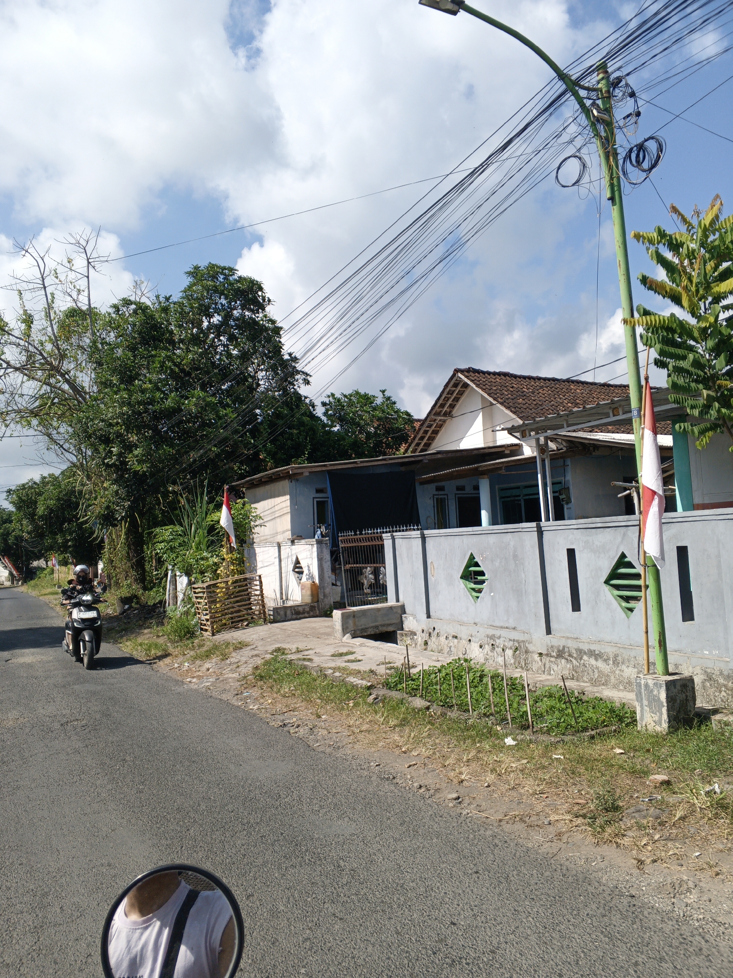 The image depicts a street scene in Padang, Indonesia, on a clear day with scattered clouds. A person is riding a motorcycle down a paved road, viewed from a first-person perspective, as indicated by the reflection of the rider in the motorcycle's mirror. The rider is wearing a helmet and a white t-shirt. To the right of the road, a gray concrete wall runs along the property line of a house. The house has a brown tiled roof and light blue walls. A green lamppost with a street light is situated near the wall, adorned with a vertical red and white flag, likely the Indonesian flag, attached to it. There are numerous electrical wires suspended from the lamppost and extending across the frame. Lush green trees line the left side of the road and the background, with some bare branches visible on one of the trees. A small patch of cultivated plants is visible next to the wall. A metal gate is present in front of the house. The overall scene suggests a residential area in a tropical climate.