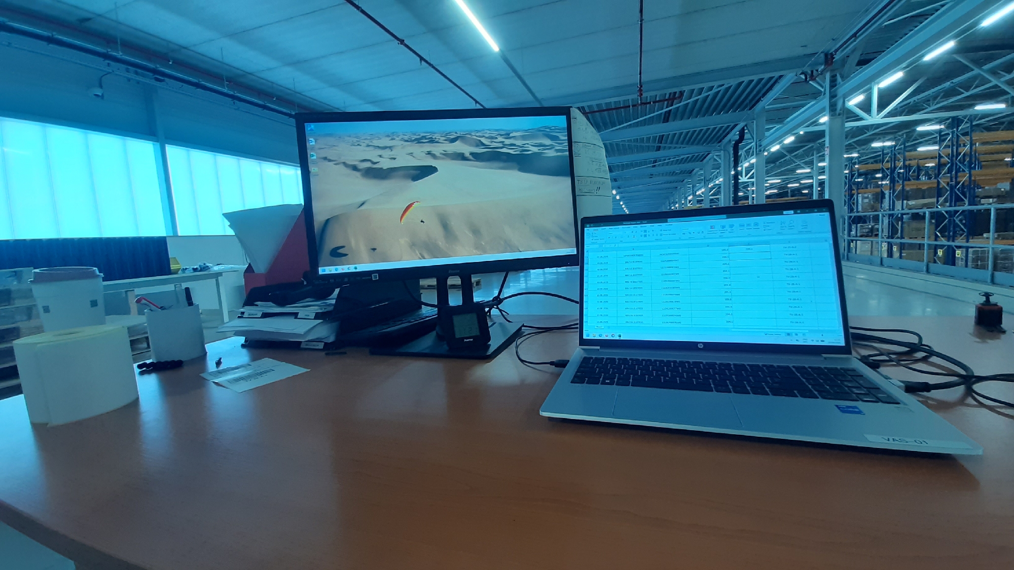 The image shows a desk in a warehouse or large industrial building, with a laptop and a monitor displaying a spreadsheet and a landscape image, respectively. The environment appears to be brightly lit, possibly by overhead fluorescent lights, and the overall color palette is dominated by cool blue tones. The desk itself is made of wood, and on it are various office supplies, including pens, papers, and a roll of what appears to be labels. In the background, rows of tall shelving units stocked with goods are visible, typical of a warehouse setting. The specific location in Hellevoetsluis, Netherlands, is not discernible from the image itself. There are no people visible in the frame.