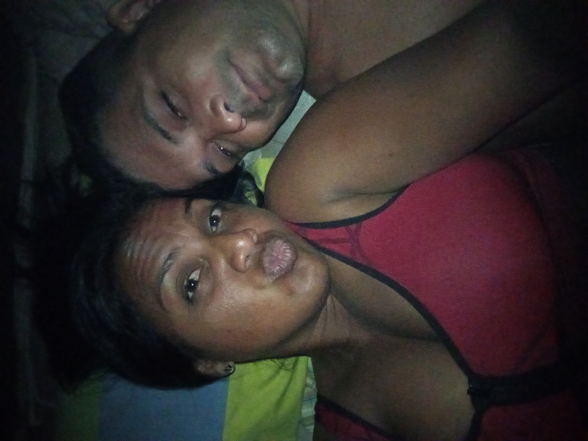 A close-up, intimate photograph shows a man and a woman lying down together, seemingly in a bed. The man is positioned behind the woman, with his face partially visible as he rests his head. His eyes are closed. The woman is lying on her side, facing the camera. She is wearing a red top with black straps and has an earring on her left ear. Her lips are puckered in a kissing expression. The bedding visible is a mix of colors, with blue, yellow, and green. The overall lighting suggests a low-light environment. The location is unknown.