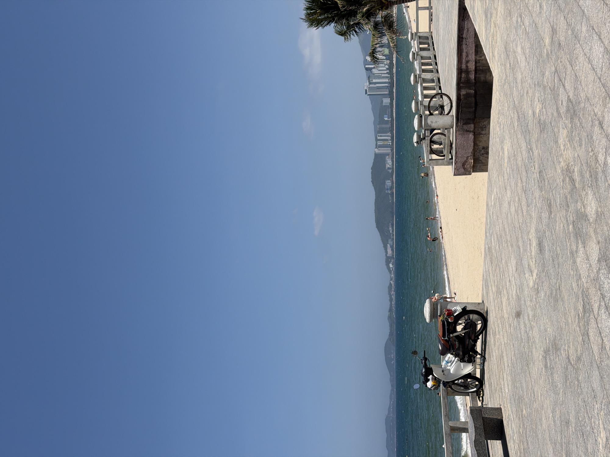 The image depicts a sunny coastal scene in Nha Trang Ward, Vietnam, viewed from a slightly elevated paved promenade. In the immediate foreground, a grey tiled surface extends across the lower portion of the frame. To the left, a grey concrete bench is visible. A white and brown classic-style scooter, possibly a Honda Super Cub, is parked on the paved area, facing left, with a front basket and a brown seat. On the right, a concrete balustrade or railing features two black bicycle wheels mounted as decorative elements.
Beyond the promenade, a light sandy beach slopes down to the turquoise-blue sea, which exhibits moderate waves. Several small, indistinct figures are visible on the beach and wading or swimming in the water. In the mid-distance across the water, verdant islands or landforms are present. The background features a vibrant city skyline dominated by numerous high-rise buildings along the coast, nestled against a range of green mountains. A portion of a palm tree is visible on the far right. The sky above is a clear, bright blue with minimal cloud cover, suggesting fair weather.