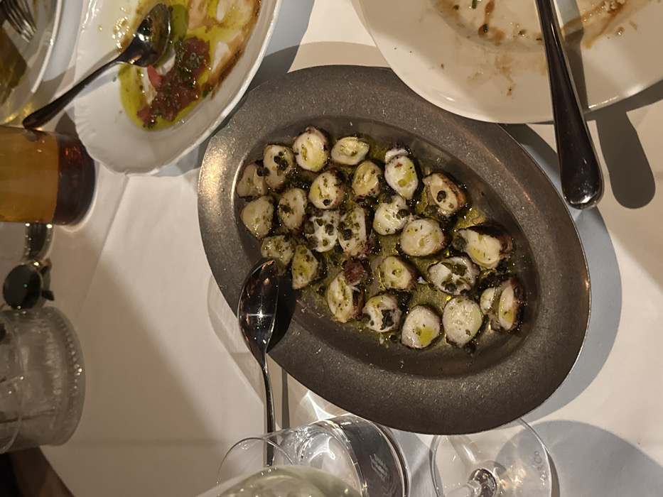 The scene depicts a restaurant table in the city of Unknown, Unknown, covered by a white tablecloth, under dim, ambient lighting. The primary subject is an oval, dark metal serving plate positioned centrally, containing approximately 25 sliced pieces of cooked octopus, thoroughly coated in olive oil and garnished with green chopped herbs and small, dark capers. A large silver serving spoon rests horizontally across the top left edge of this plate.
To the upper right, a white, scalloped-edge oval plate contains remnants of a dish featuring a red, textured sauce (likely tomato-based) and a pool of olive oil, suggesting a partially consumed carpaccio or similar appetizer. A silver spoon is positioned within this plate. Below this, to the lower right, a segment of another white plate with food residue is visible, with a silver spoon lying beside it.
To the left of the main octopus dish, a clear stemmed wine glass, bearing a "Tiffany & Co." inscription on its base, is partially filled with a pale yellow liquid, consistent with white wine. Above this, a clear glass tumbler with a textured, ribbed base is present. Further in the background, on the white tablecloth, a dark brown glass bottle and a pair of dark sunglasses are visible. No individuals are depicted in the image.