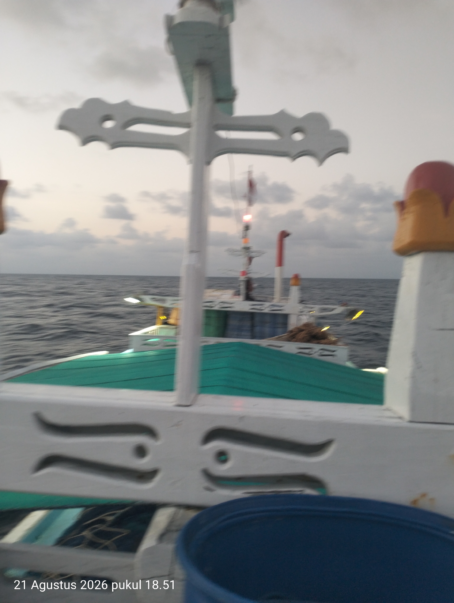 The image is a slightly blurry, low-angle, close-up shot taken from the deck of a boat. In the foreground, the white railing of the boat is visible, featuring decorative carved white elements that resemble stylized eyes or abstract shapes. A large, dark blue plastic container is in the lower right corner of the frame. Behind the railing, the turquoise-green covering of the boat's deck is visible, extending towards the horizon.
In the mid-ground, another boat or part of the same vessel is seen, with a white superstructure and green accents. There are visible lights and some brown, fibrous material, possibly fishing nets or cargo, piled on the deck. A tall mast with red and white sections and a red light is prominent, suggesting this is a fishing vessel.
The background consists of a wide expanse of greyish-blue ocean with moderate waves, stretching to a hazy horizon. The sky above is overcast with muted grey clouds, indicating either early morning, late evening, or poor weather conditions.
The timestamp at the bottom left corner reads "21 Agustus 2026 pukul 18.51," which translates to "August 21, 2026, 18:51." This timestamp indicates the image was taken in Indonesia, specifically at 6:51 PM on August 21, 2026. The exact location within Indonesia is not specified in the image, but the context of a boat at sea suggests a coastal or offshore environment. There are no people clearly visible in the image.