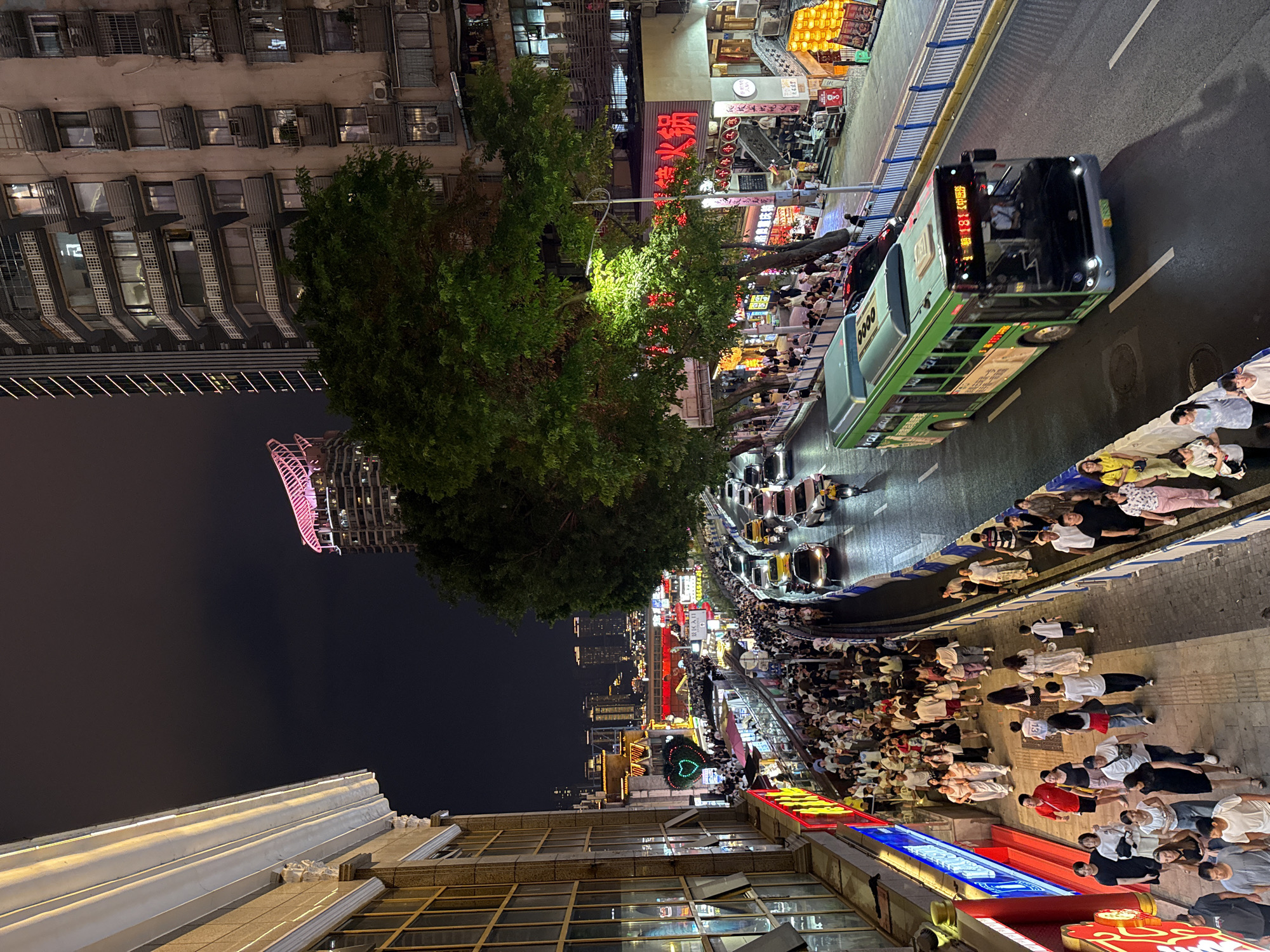 This image depicts a bustling street scene at night in the Yuzhong District of Chongqing, China. A green bus is traveling down a multi-lane road, which is lined with pedestrians walking on elevated sidewalks. Buildings with illuminated signs and windows flank the street, and a large tree with green foliage stands prominently on the right side of the image. The sky is dark, suggesting it is nighttime. The architecture appears to be a mix of modern high-rise buildings and older structures. The sidewalks are crowded with people, many of whom are casually dressed, indicating a lively urban environment. Various vehicles, including cars and motorcycles, are also present on the road. The overall impression is one of a vibrant and active city at night.