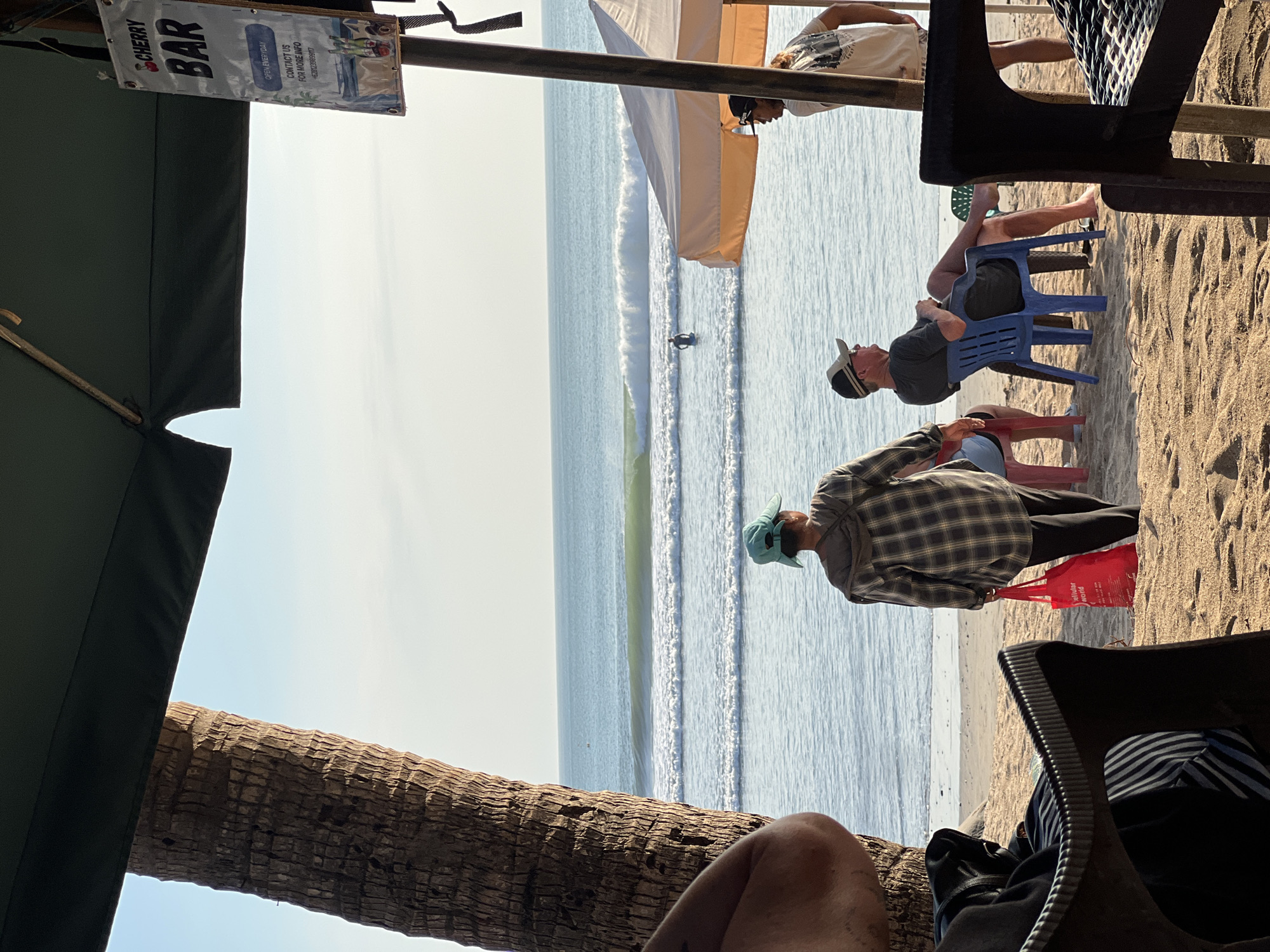 The image depicts a beach scene in Kuta, Indonesia, on a bright day. In the foreground, a person's arm is visible on the left, and a black plastic chair with striped cushions is in the lower left corner. Further back, a person wearing a plaid shirt and a blue hat is walking towards the ocean, carrying a red bag. Two men are seated on chairs facing the sea; one is wearing a hat and dark t-shirt, and the other is partially visible. A surf spot with waves is in the midground, with a surfer in the water. A tent-like structure is visible on the right, and a sign for "CHERRY BAR" hangs from a pole. A palm tree trunk is to the left, framing the scene. The ocean stretches to the horizon under a clear sky.