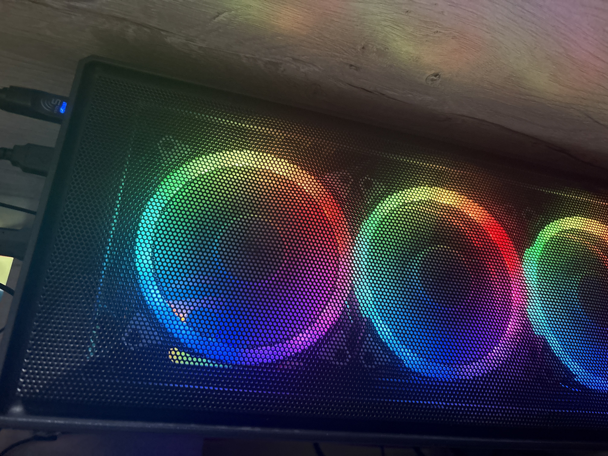 The image shows a computer tower with three RGB fans visible through a mesh front panel. The fans are illuminated with a gradient of colors, including blue, purple, green, yellow, and red. The computer is positioned in front of a wooden surface, possibly a wall or desk. There are some cables connected to the top of the computer, one of which has a blue indicator light. The lighting suggests the room is dimly lit, with the RGB lighting of the computer being the primary light source. The image does not provide any information about the city of Tulinskiy, Russia.