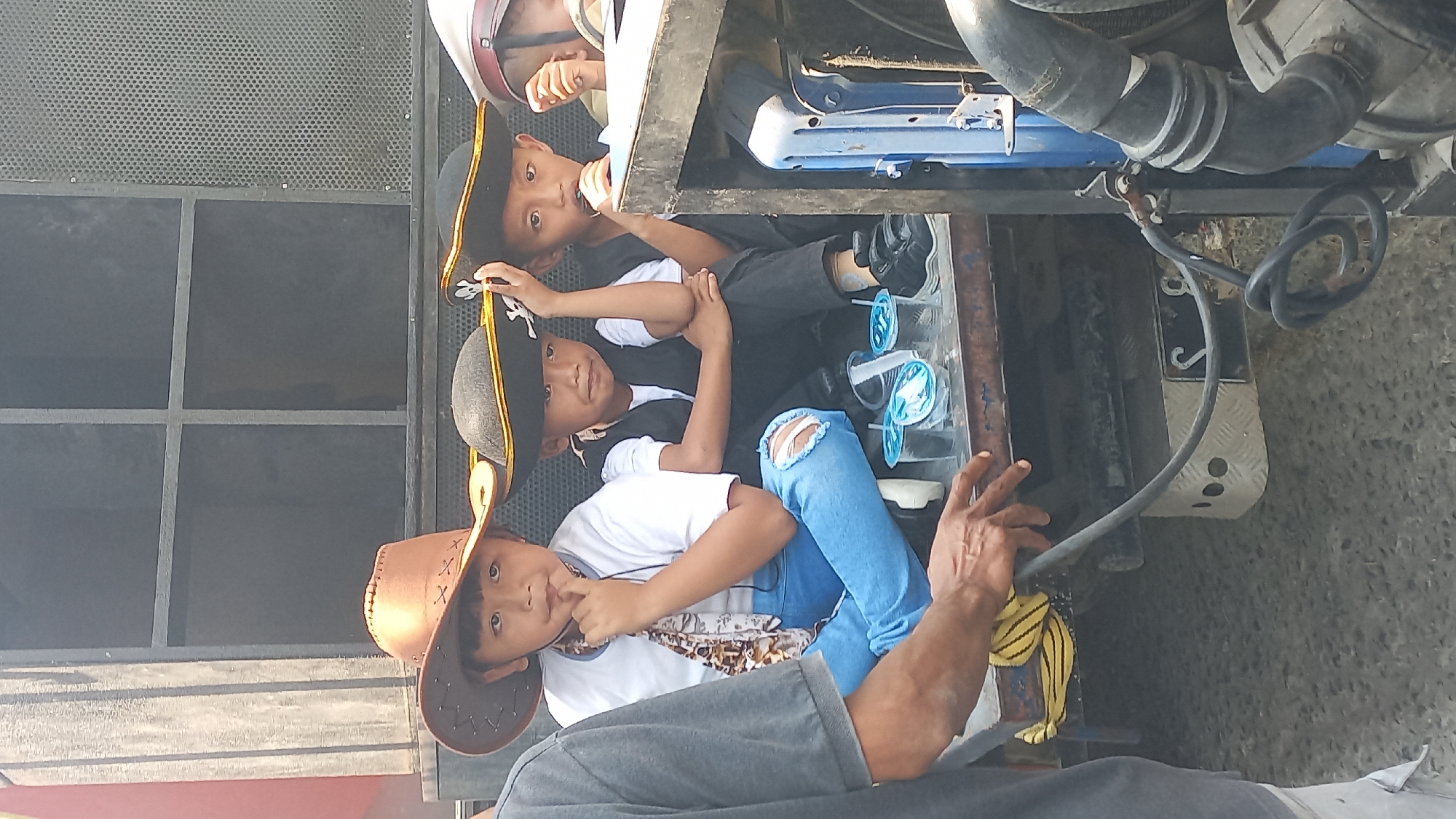 In Kedungmegarih, Indonesia, three young boys are seated on the back of a truck. The boy on the left wears a brown cowboy hat, a white t-shirt, and blue jeans with ripped knees. He has a patterned scarf around his neck and rests his chin on his hand. The boy in the middle wears a black pirate hat, a black vest over a white t-shirt, and black pants. He has his left hand on his hip and his right hand touching his chin. The boy on the right also wears a black pirate hat, a black vest over a white t-shirt, and black pants. He holds his right hand up to his mouth. In front of the boys are several clear plastic cups with blue liquid. The truck is parked on a grey, textured surface. Behind the boys is a large, black, grid-like structure, possibly a speaker system. To the right of the boys is a blue metallic object and industrial hoses. A person's arm and hand, wearing a grey shirt, are visible on the left side of the frame, interacting with a yellow and black striped cloth on the truck. The overall scene suggests a festive or event-related setting.