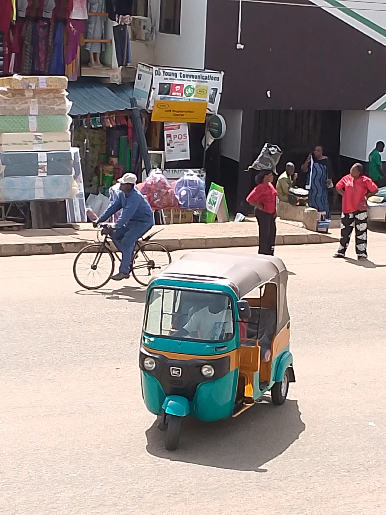 In Bukuru, Nigeria, a bustling street scene unfolds under what appears to be a bright, sunny day, suggested by the strong shadows cast by objects and people. The foreground is dominated by a teal and yellow auto-rickshaw, also known as a "keke," its passenger compartment open, revealing a hint of the driver. The rickshaw is positioned on a paved road, implying an urban or semi-urban environment.
Further back, a man in a blue jumpsuit and a white cap rides a bicycle, his form creating a distinct shadow on the road. He is moving from left to right in the frame. Behind him, a market or shop area is visible, with stacks of mattresses or bedding wrapped in plastic on the left. On the right side of the street, a group of people are gathered. One woman, wearing a black and blue patterned dress, stands and appears to be in conversation, possibly on a phone. Another woman in a red top and patterned pants stands with her hands on her hips, looking towards the viewer. A man in a red shirt and dark pants is also present, his posture suggesting he is listening or observing.
A sign above a building reads "DS Young Communications" and displays logos for "Glo" and "Airtel," indicating a telecommunications provider. Another sign nearby says "POS," suggesting a point-of-sale service. The architecture of the buildings suggests a commercial area, with awnings and storefronts. The overall impression is one of everyday activity in a lively Nigerian marketplace, with a mix of transportation, commerce, and social interaction. The clothing styles and the presence of the auto-rickshaw and the bicycle point to a warm climate.