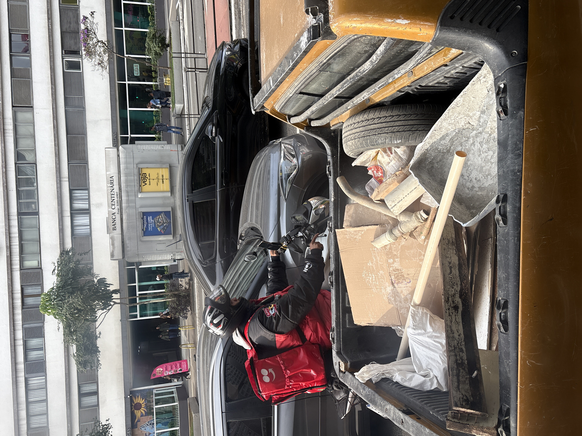 A delivery person wearing a red and black jacket and a helmet rides a motorcycle on a street in São Paulo, Brazil. They are carrying a large red insulated delivery bag on their back. The motorcycle is in the foreground, positioned to the left of the frame, and appears to be stopped in traffic. Behind the motorcycle, there are several cars, including a black sedan and a gray sedan, also stopped. In the background, a building with multiple windows and blinds is visible, with a banner that reads "BANCA CENTENÁRIA" and "VEJA." There are people walking on the sidewalk in front of the building. The street appears to be an urban environment with paved roads and sidewalks. The delivery person's motorcycle is partially obscured by the bed of an orange pickup truck, which is filled with construction debris, including cardboard boxes, pipes, and a shovel. The lighting suggests it is daytime.