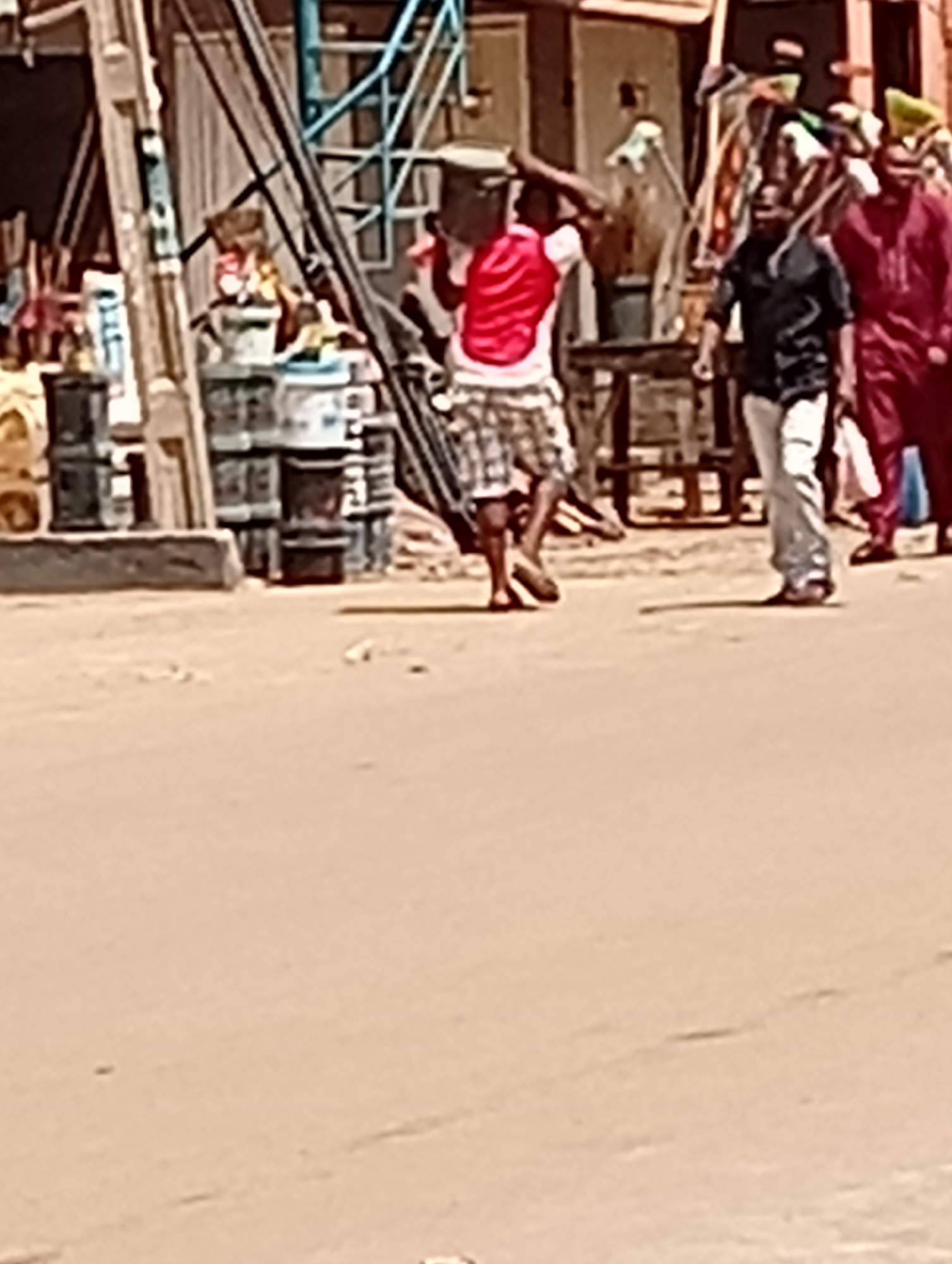 The image depicts a street scene in Maiburiji, Nigeria. In the foreground is a tan-colored street. A man wearing a red and white shirt and plaid shorts carries an object on his head. He walks along the side of the road. To his right is another man wearing a black shirt and light-colored pants. Further to the right are two men, one wearing a red outfit and another whose outfit is partially visible. In the background are stacks of barrels and several structures. Overhead utility lines are also present.