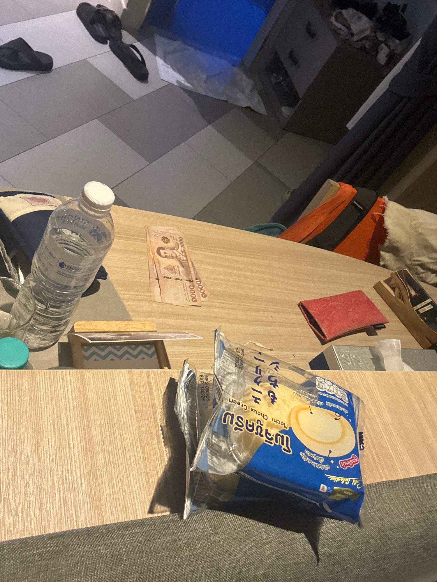 In Karon, Thailand, a wooden table surface holds several items. A clear plastic water bottle with a white cap stands upright. Two Thai baht bills, one appearing to be a 1000 baht note, lie near the bottle. A small red cloth or wallet is placed to the right of the bills. In the foreground, a plastic-wrapped food item, possibly a mochi choux cream snack, is visible. The background shows a tiled floor with two black flip-flops. Further in the background, there is a dark piece of furniture with some items on it, and a glimpse of orange fabric.