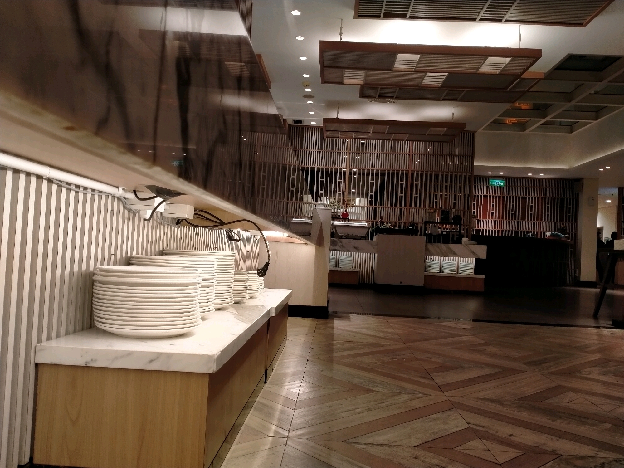 The image depicts a well-lit buffet area, likely in a hotel or restaurant. In the foreground, several stacks of white plates are neatly arranged on a marble-topped counter. The counter is supported by a wooden cabinet. Behind the plates, vertical striped wallpaper is visible. The ceiling features modern wooden paneling and recessed lighting. Further back, a decorative wooden screen divides the space. An "EXIT" sign with a green arrow is prominently displayed on the screen. The floor is made of polished wood with a geometric pattern. The overall ambiance suggests a clean and organized dining establishment. The image is a photograph taken in the city of Batu, Indonesia, specifically at the Grand Mercure Malang Mirama hotel.
