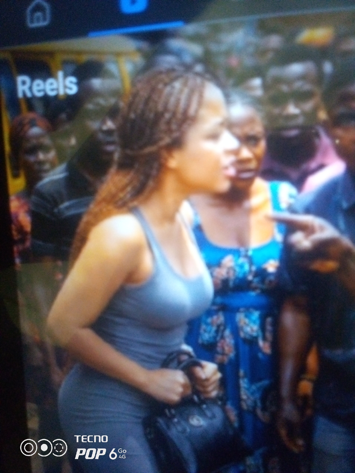 A crowded street scene, likely in Howayi, Nigeria, features several individuals. In the foreground, a woman with braided hair is wearing a grey tank top and a dark purse is visible. Behind her, another woman is seen in a blue and floral patterned dress. Several other people, mostly men, are present in the background, some looking towards the camera. The environment appears to be outdoors, with a yellow vehicle partially visible in the upper left quadrant. The overall lighting suggests it may be daytime. A logo that reads "TECNO POP 6 4G" is present in the lower left corner, indicating the image was likely captured by a Tecno phone.