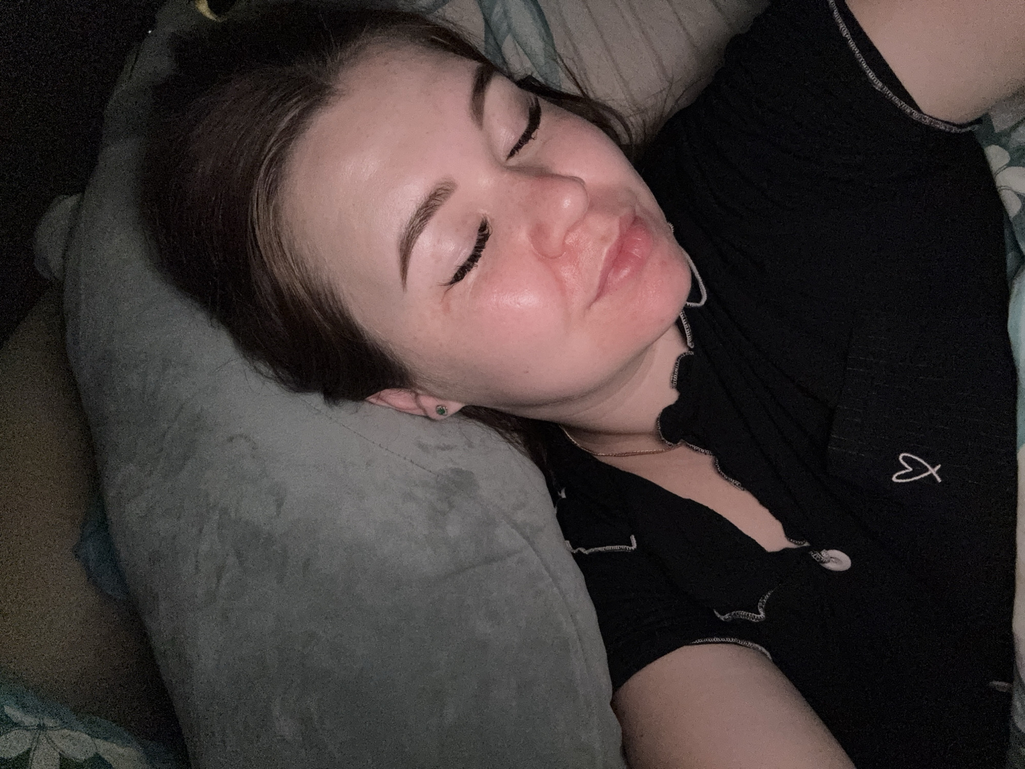 A young woman is lying on her side with her eyes closed, appearing to be asleep or resting. She is wearing a black t-shirt with white stitching around the neckline and sleeves, and a small embroidered heart on the chest. She has brown hair and is wearing a small green earring. The setting appears to be indoors, possibly a bedroom, with a pillow behind her head and a blanket or comforter visible. The lighting is dim, suggesting it might be nighttime or a dimly lit room. There is no indication of the location being in the city of Chaikovsky, Russia.
