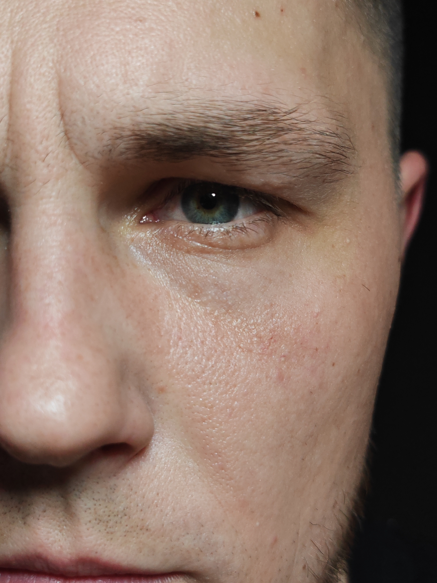 Close-up of a man's face, cropped on the left and right sides. The man has light skin, short dark hair, a prominent brow, and green-blue eyes. Fine wrinkles are visible on the forehead and around the eye. Sparse facial hair is present near the lip and along the jawline. The background is dark and out of focus. There is no indication of the location of Hrodna, Belarus.