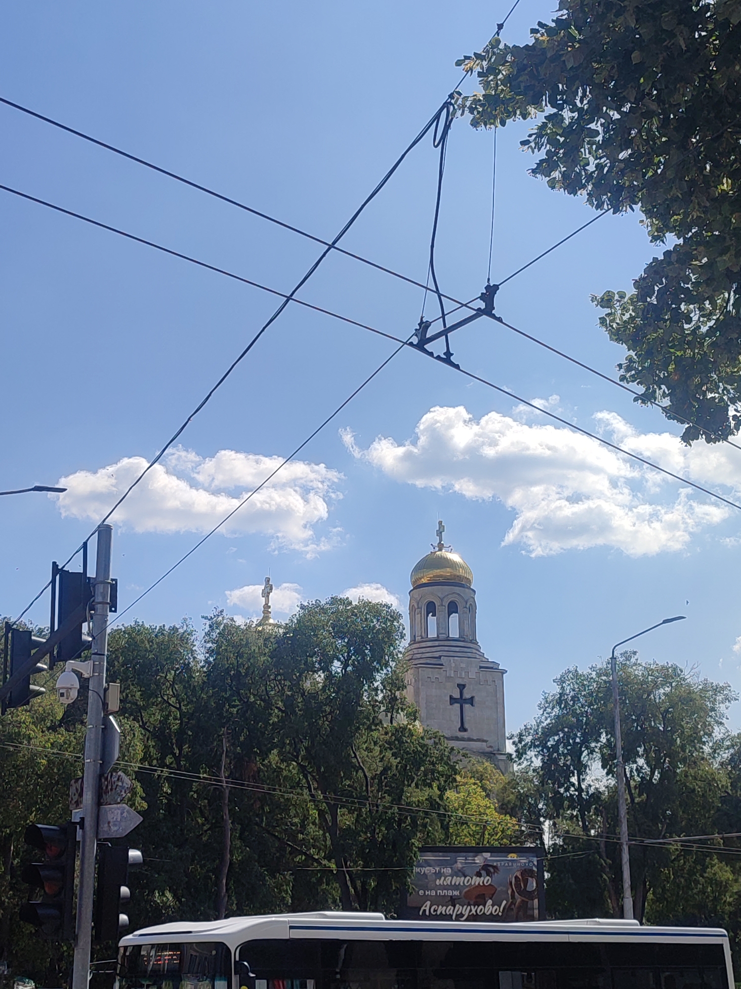 The image shows a bright, sunny day in Varna, Bulgaria, with a blue sky and fluffy white clouds. Several overhead power lines crisscross the frame, with lush green trees framing the scene. In the background, the distinctive golden dome of the Dormition of the Mother of God Cathedral, also known as the Varna Cathedral, is visible, topped with a cross. Next to the cathedral, a tall bell tower with a large cross on its facade stands prominently.
In the foreground, a white bus is partially visible at the bottom of the frame. To the left, a traffic light pole with multiple lights and a security camera is present. A street sign points to the right. Below the cathedral, a billboard with text in Bulgarian and images of people enjoying summer activities is displayed. The overall impression is of a vibrant urban setting within the city of Varna.