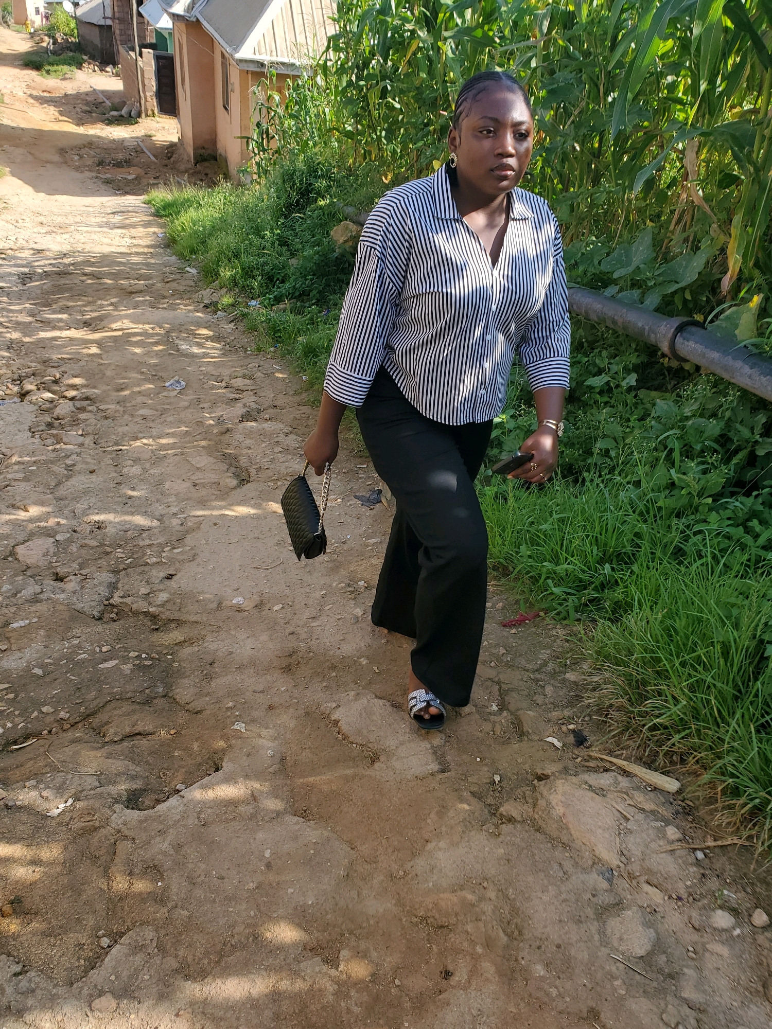 A young Black woman walks on a dirt road in a rural area that appears to be the city of Jos, Nigeria. She is wearing a black and white striped shirt, black pants, and silver sandals. She is carrying a black purse in her right hand and a mobile phone in her left hand. The road is unpaved and has some potholes. On the right side of the road, there are tall green plants, likely corn stalks, and thick green grass. On the left side of the road, there are a few buildings with corrugated metal roofs, suggesting a residential area. The woman is looking to her left and appears to be walking towards the viewer. The lighting suggests it is daytime, with sunlight casting shadows on the road.