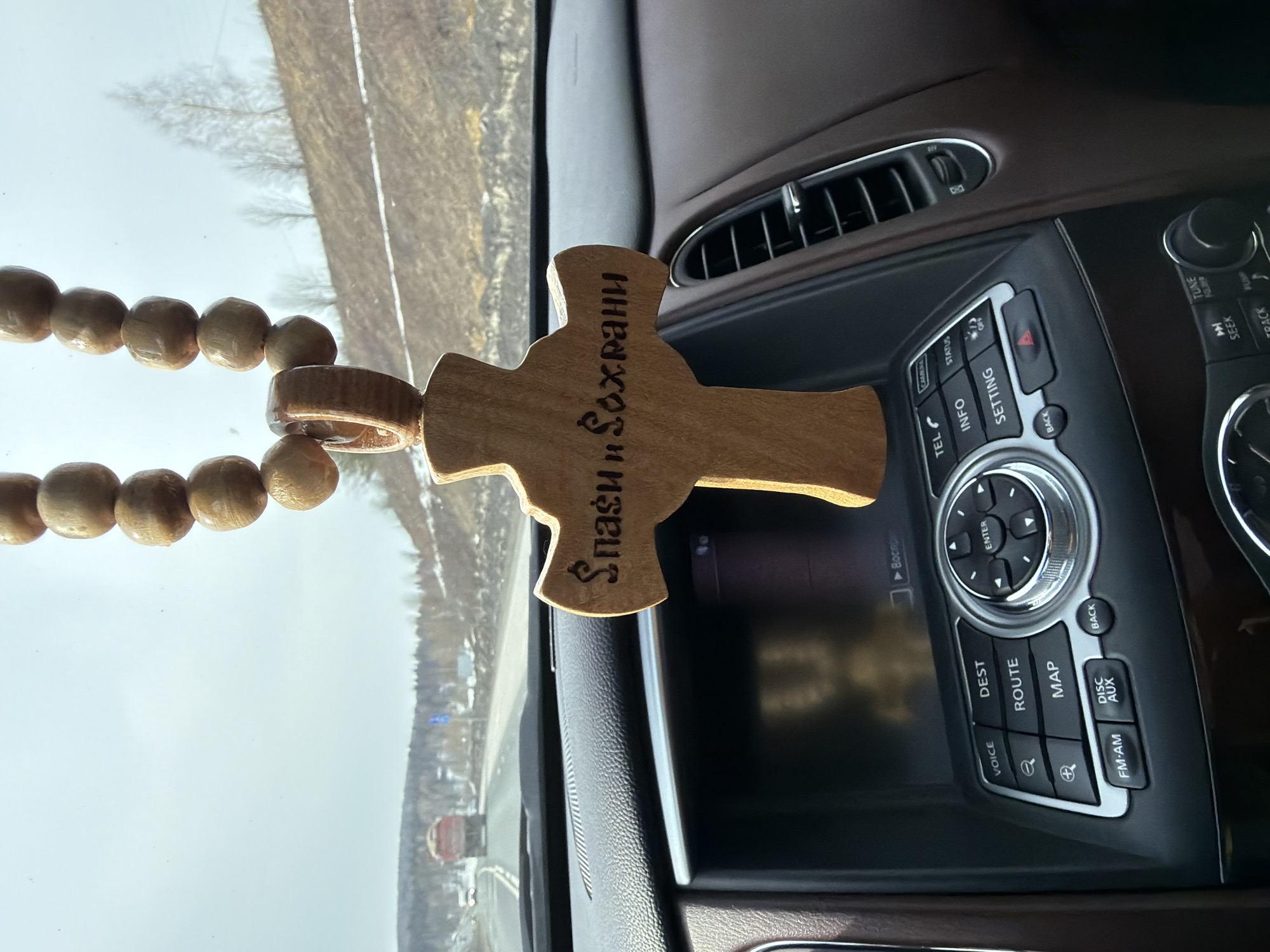 The image depicts a view from the interior of a vehicle on a paved road in Ust'-Shalashnaya, Russia, during what appears to be late winter or early spring. A string of light brown wooden beads, resembling a rosary, hangs from the rearview mirror area. Attached at the bottom is a light brown wooden cross, inscribed with the Cyrillic text "Спаси и Сохрани" (Save and Protect). The vehicle's dark gray dashboard is visible in the foreground, featuring an infotainment system with a dark screen and various control buttons, including "VOICE," "DEST," "ROUTE," "MAP," and a central rotary dial. Brown leather-like trim is visible on the passenger side dashboard. Through the windshield, a two-lane asphalt road extends into the distance under an overcast sky. A red dump truck is visible ahead in the right lane, traveling in the same direction. The surrounding environment includes forested hills on the left and a barren, brownish hillside with patches of snow and sparse deciduous trees on the right. A blue road sign is partially visible on the right side of the road.
