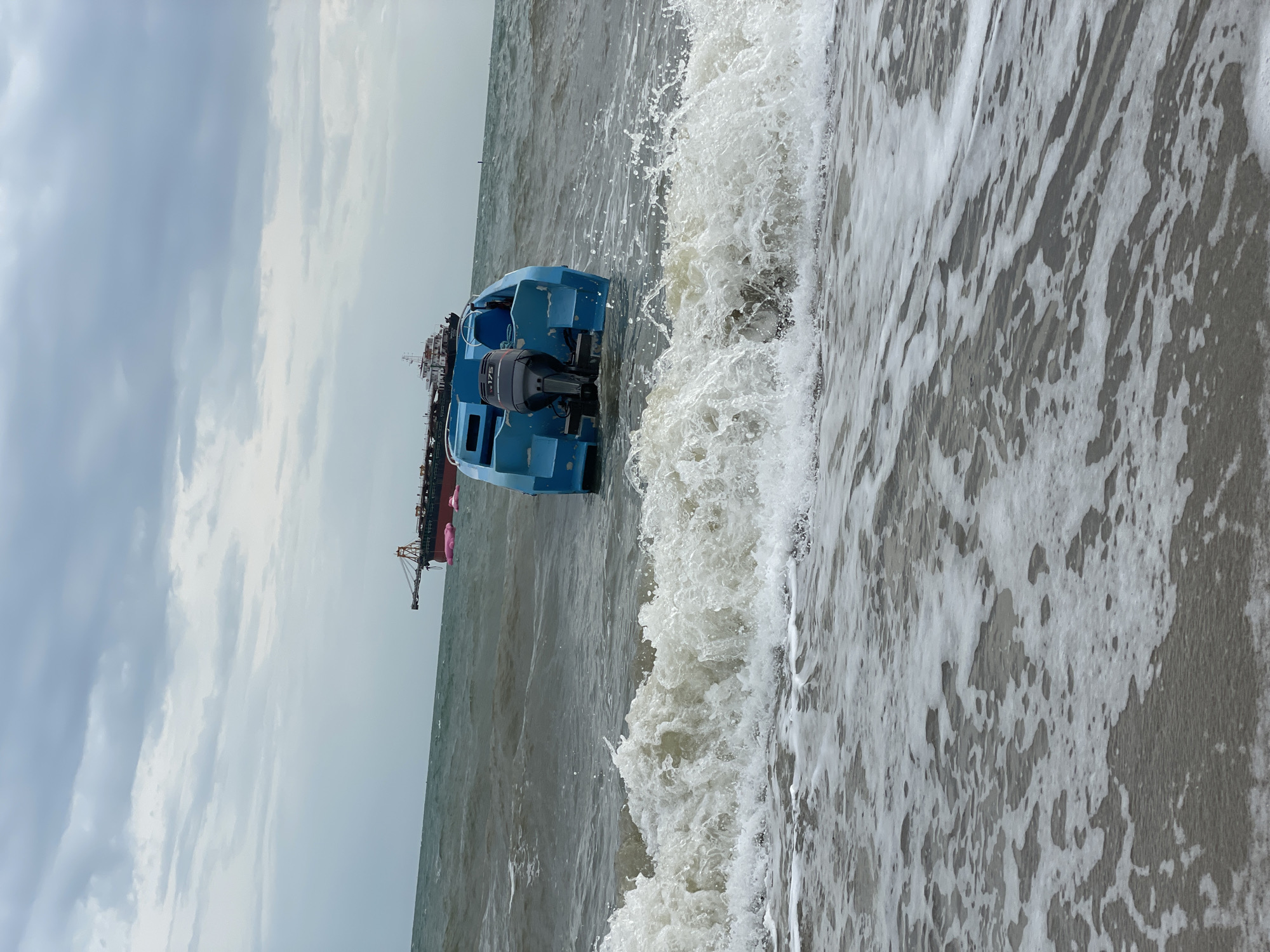 In Pattaya City, Thailand, a blue motorboat with an outboard motor is partially submerged in choppy ocean water near the shore. The boat is positioned facing away from the camera. In the background, two large cargo ships are visible on the horizon under a cloudy sky. Waves are breaking in the foreground, creating foam that washes onto the sandy beach. The scene appears to depict a marine environment with commercial shipping activity and a small recreational boat in distress.