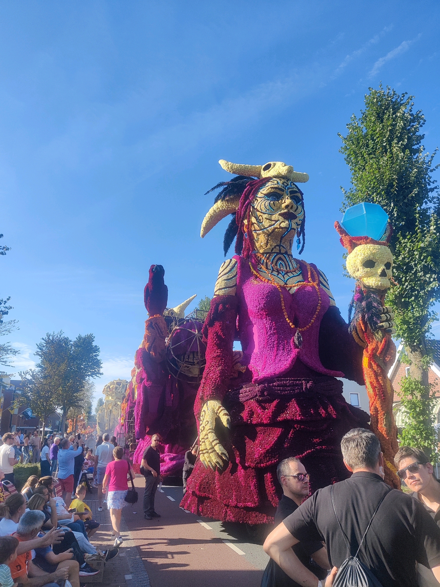 The image captures a vibrant street parade in Zundert, Netherlands, likely during the annual Bloemencorso Zundert flower parade. The central focus is a colossal, meticulously crafted float depicting a large figure adorned with a skull headdress and elaborate floral decorations in shades of yellow, purple, and red. The figure holds a staff topped with a skull and a blue, faceted object. Behind this main float, other floral floats and structures are visible, stretching down the street. A crowd of spectators lines the street, some seated on benches and others standing. Many are looking towards the floats, with some holding up cameras to capture the spectacle. The environment is a sunlit day with a clear blue sky and green trees flanking the street. The parade appears to be moving along a paved road, with buildings and houses visible in the background, indicating a residential or town center setting. The people in the image are casually dressed, suggesting a festive, outdoor event.