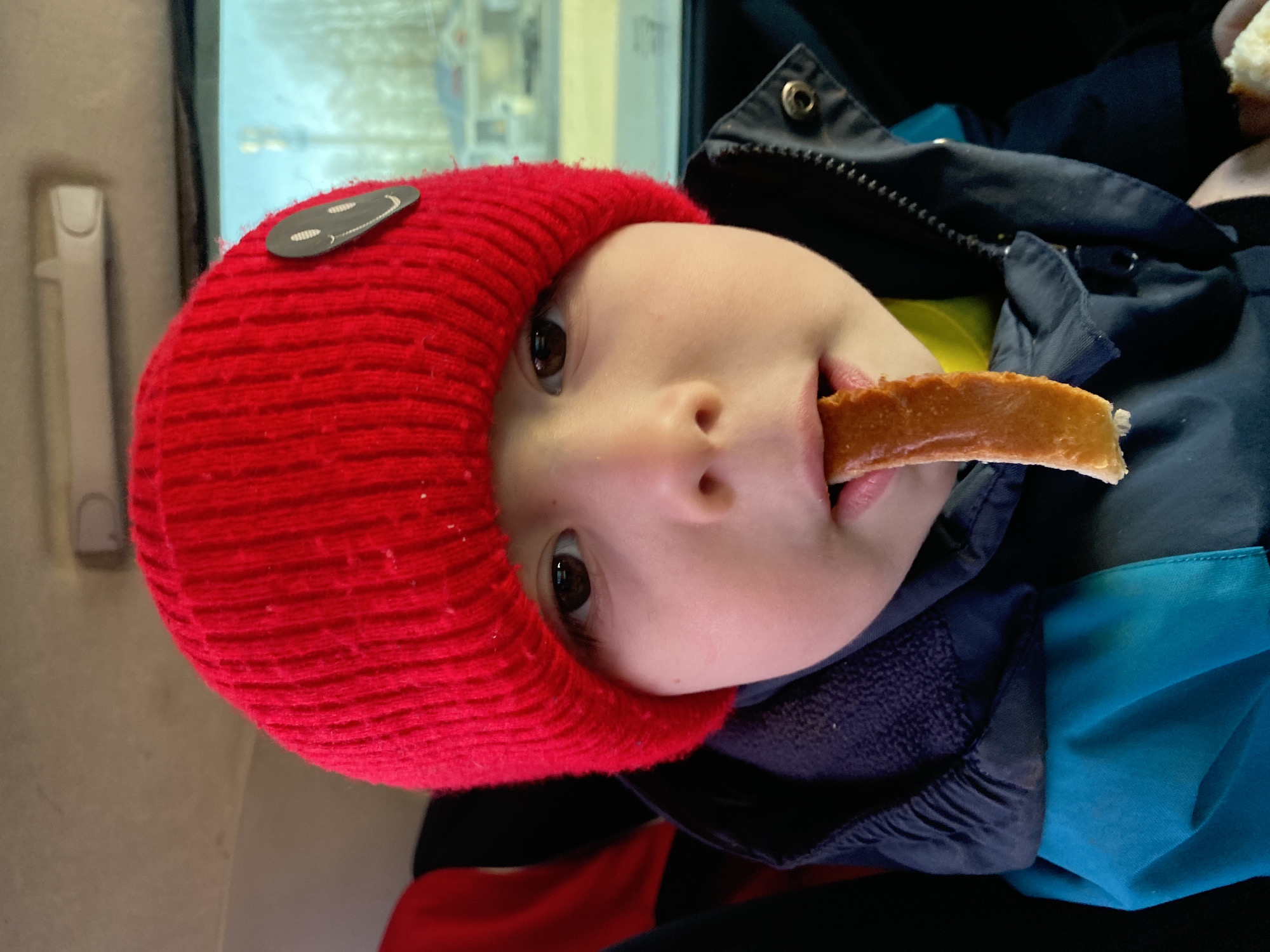 A baby wearing a red knitted hat with a black oval patch with a smiley face stitched onto it, is eating a piece of bread. The baby is wearing a dark blue and turquoise jacket. The baby is in Chaikovsky, Russia, based on reflections in the window, which include a Cyrillic lettered sign outside of the building.