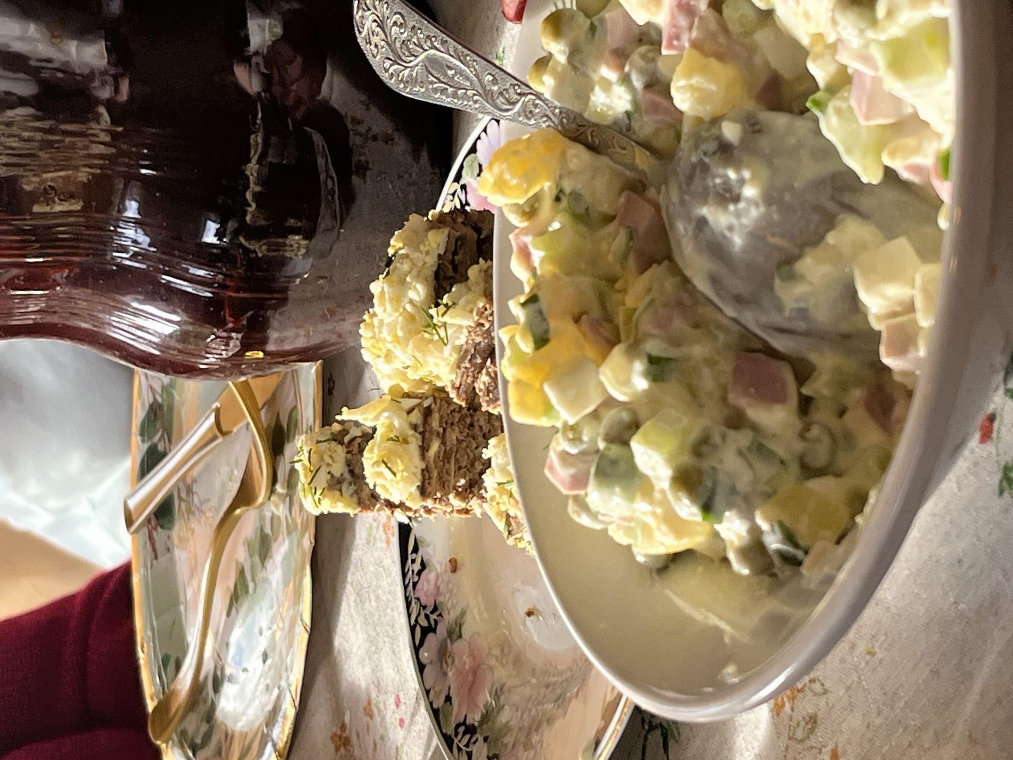 The image shows a table setting with food. A bowl filled with Olivier salad, mixed with potatoes, peas, ham, cucumbers, and mayonnaise, sits prominently in the foreground. A silver spoon is nestled within the salad. Next to the bowl, there's a plate holding a sliced meatloaf, garnished with chopped eggs and herbs. A dark-colored beverage in a glass bottle is positioned behind the food. A decorative plate with a golden handle is visible near the bottle. The table is covered with a patterned tablecloth. Based on the metadata, the location is Фоки, Russia.