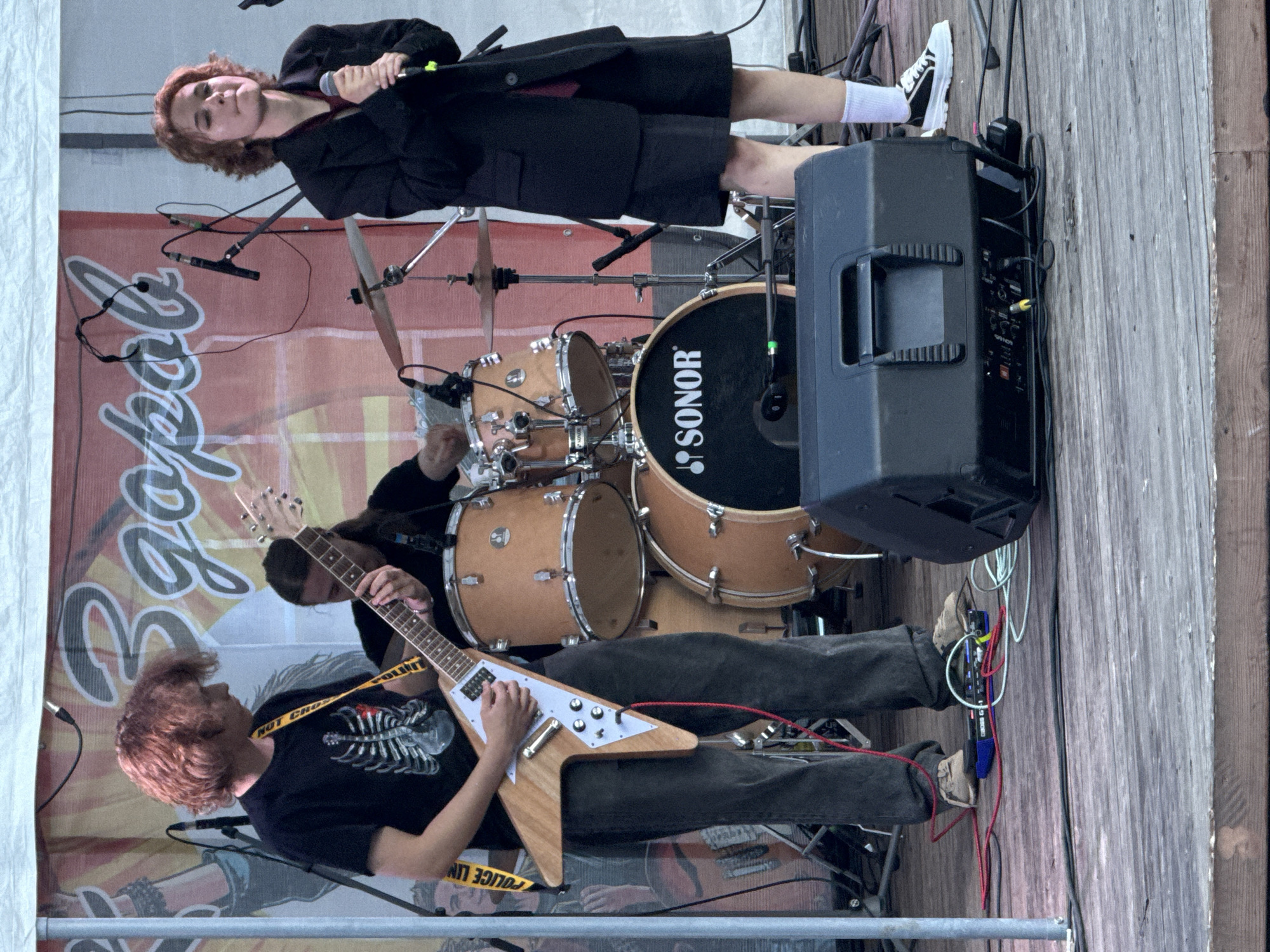 An outdoor stage performance is captured from a medium shot, with a band playing on a wooden platform under a white canopy. In the foreground, a large black speaker is positioned to the right, with various cables and electronic equipment, including a blue BOSS GT-1 pedalboard, visible on the stage floor.
Three individuals are present. On the left, a person with reddish, curly hair plays a light-colored V-shaped electric guitar. They are wearing a black t-shirt with a graphic of a ribcage and dark pants. A yellow strap with "POLICE LINE DO NOT CROSS" printed on it is visible across their shoulder.
In the center background, a drummer with long dark hair is seated behind a drum kit. The drums are light brown with black accents and a prominent "SONOR" logo on the bass drum.
To the right, a person with short reddish hair and a dark blazer over a dark shirt is holding a microphone and singing. They are wearing dark shorts and white socks with black and white sneakers.
The backdrop behind the band features a large banner with Cyrillic lettering in white and red. The overall scene suggests a casual, live music event, potentially part of a festival or local gathering. The presence of the "SONOR" drum brand and the specific "BOSS GT-1" pedal suggest the band is equipped for a modern performance. The image was taken in the city of Votkinsk, Russia, as indicated by the provided context.